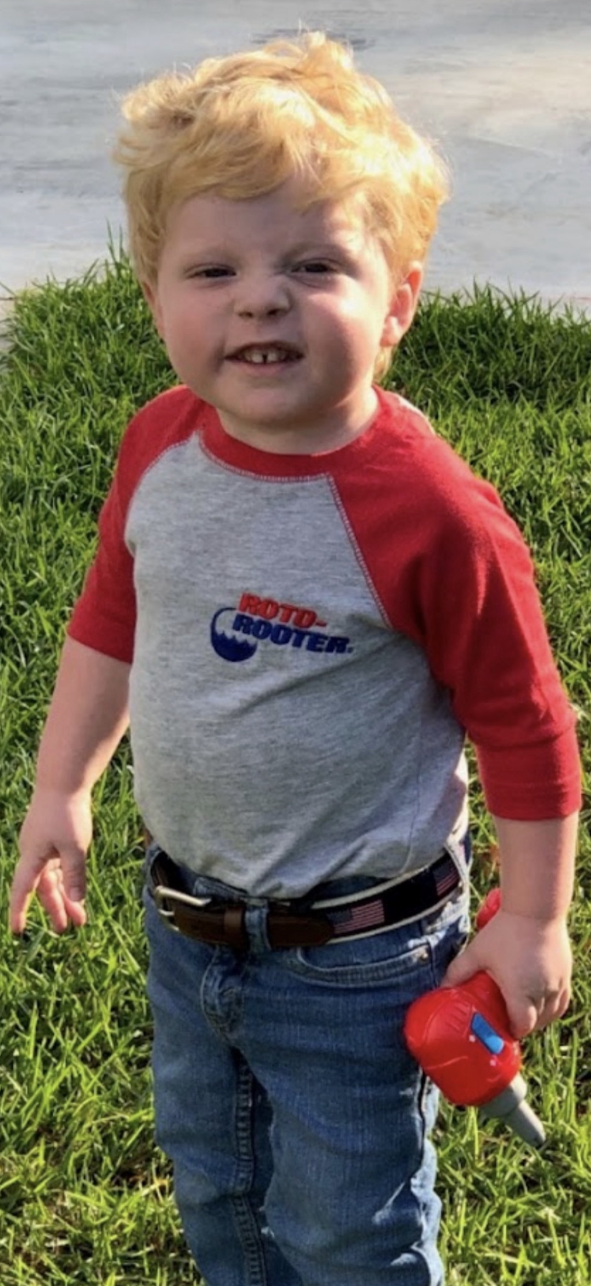 A young child standing on grass wearing a grey and red baseball shirt and jeans, holding a toy tool.
