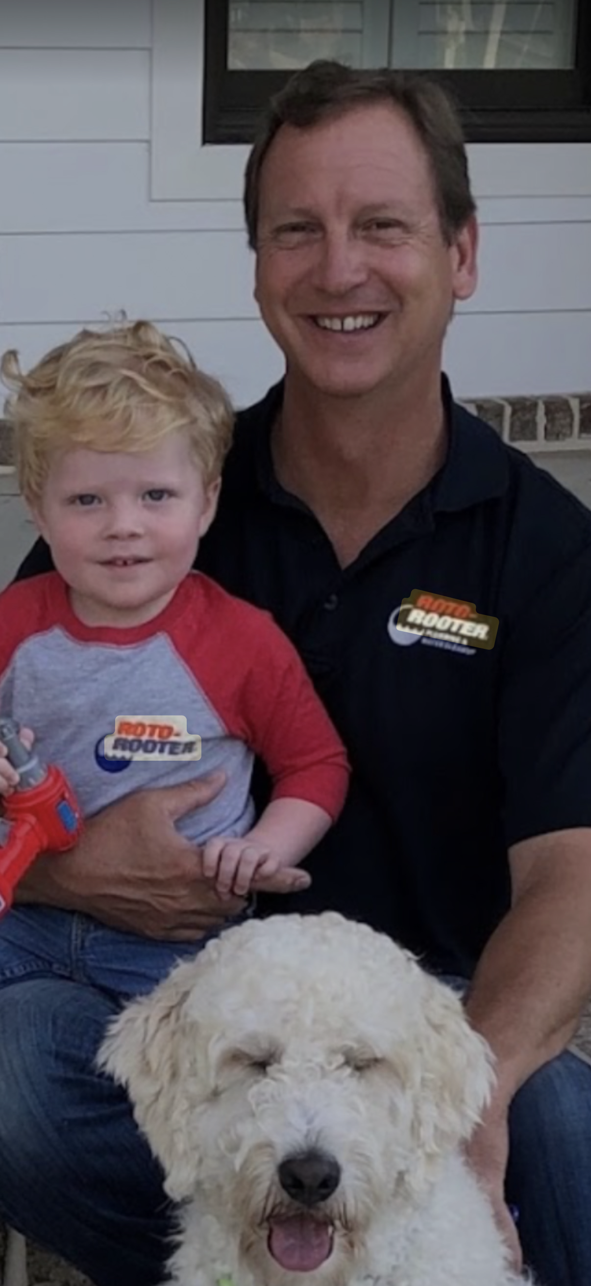 A man and a young child wearing Roto-Rooter shirts sit outdoors with a fluffy, white dog smiling at the camera.