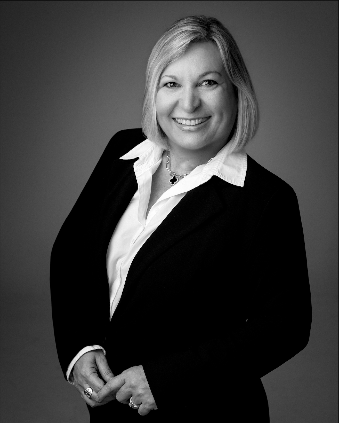 A professional headshot of a smiling person wearing a dark blazer and white collared shirt against a gray background.
