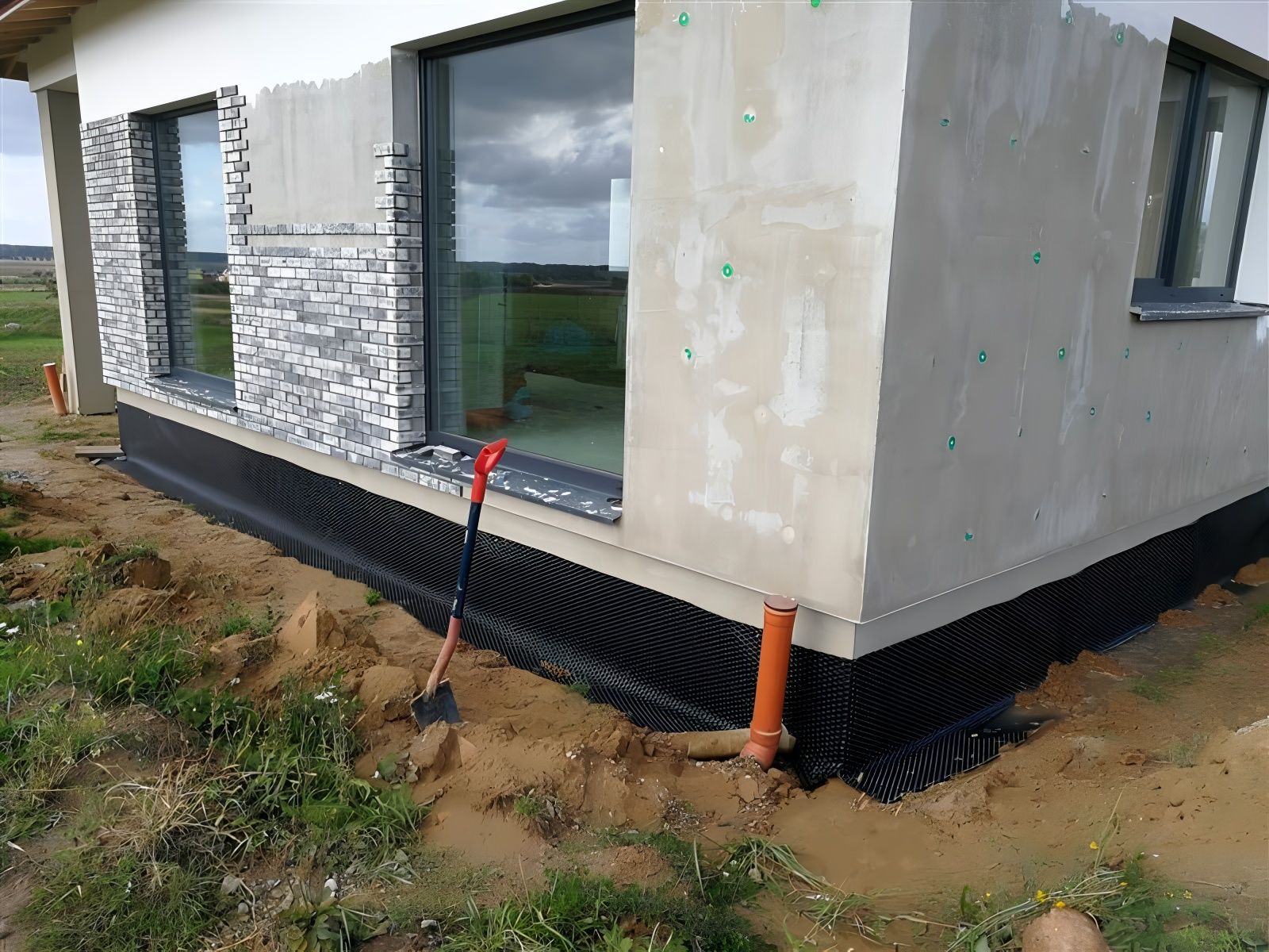 Exterior house corner under construction; black drainage membrane; dirt and tools.