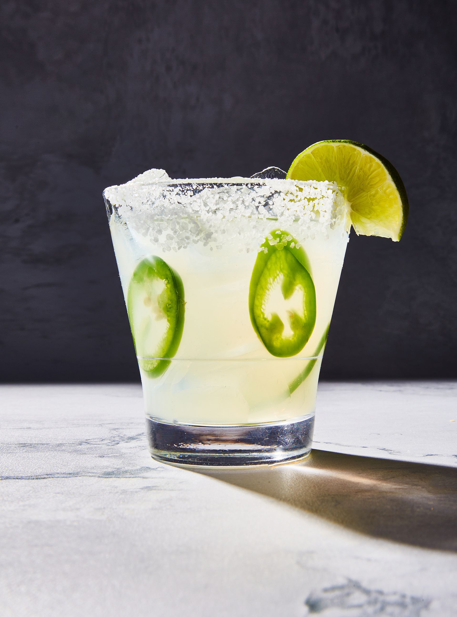 A close up of a margarita with jalapenos and a lime slice on a table.
