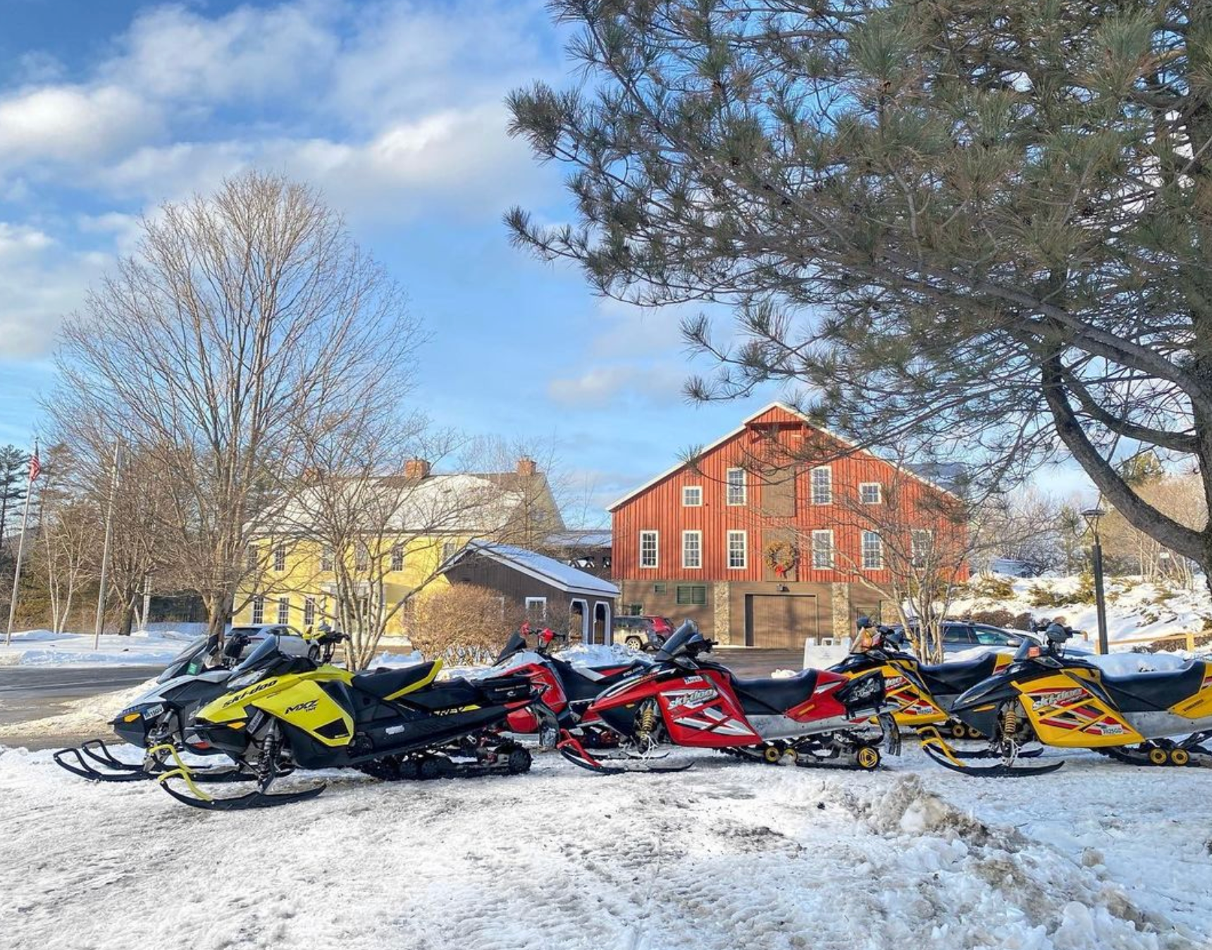 A row of snowmobiles are parked in the snow in front of a red barn.