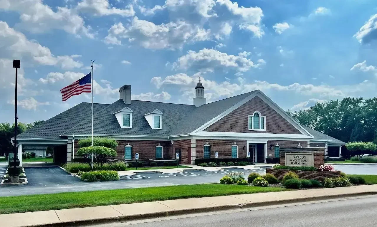 A single-story brick building with a dark shingled roof, a flagpole, and landscaping, viewed from across a road.