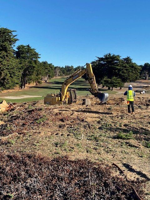 Yellow excavator clearing land on a golf course, with worker in safety vest. Blue sky, green grass.