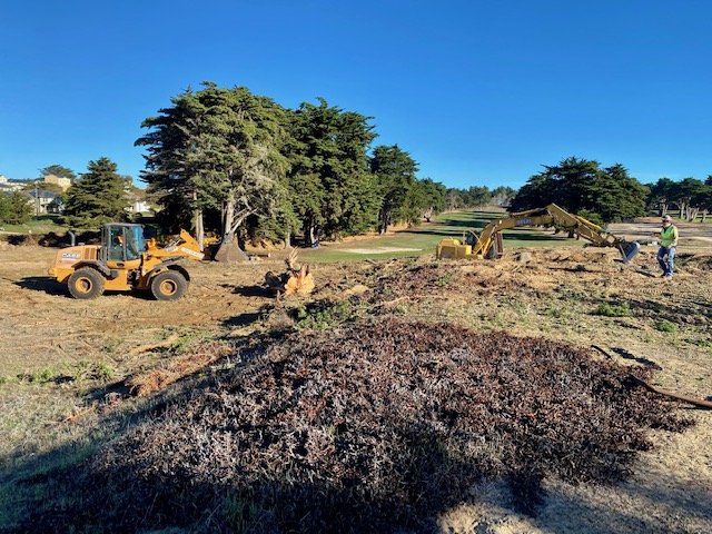 Construction on a golf course: A yellow loader and excavator with workers clearing brush and debris.