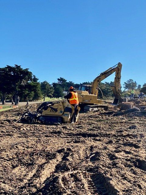 Construction site with excavator and worker in orange vest on a dirt field under a blue sky.