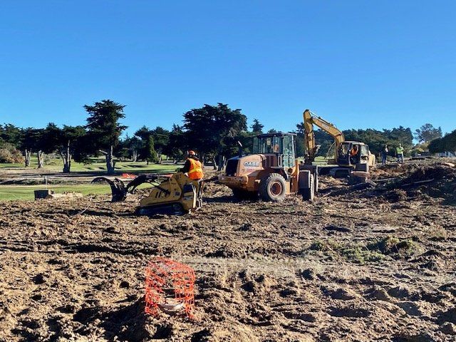 Construction site with heavy machinery like excavators and a loader on muddy ground under a blue sky.