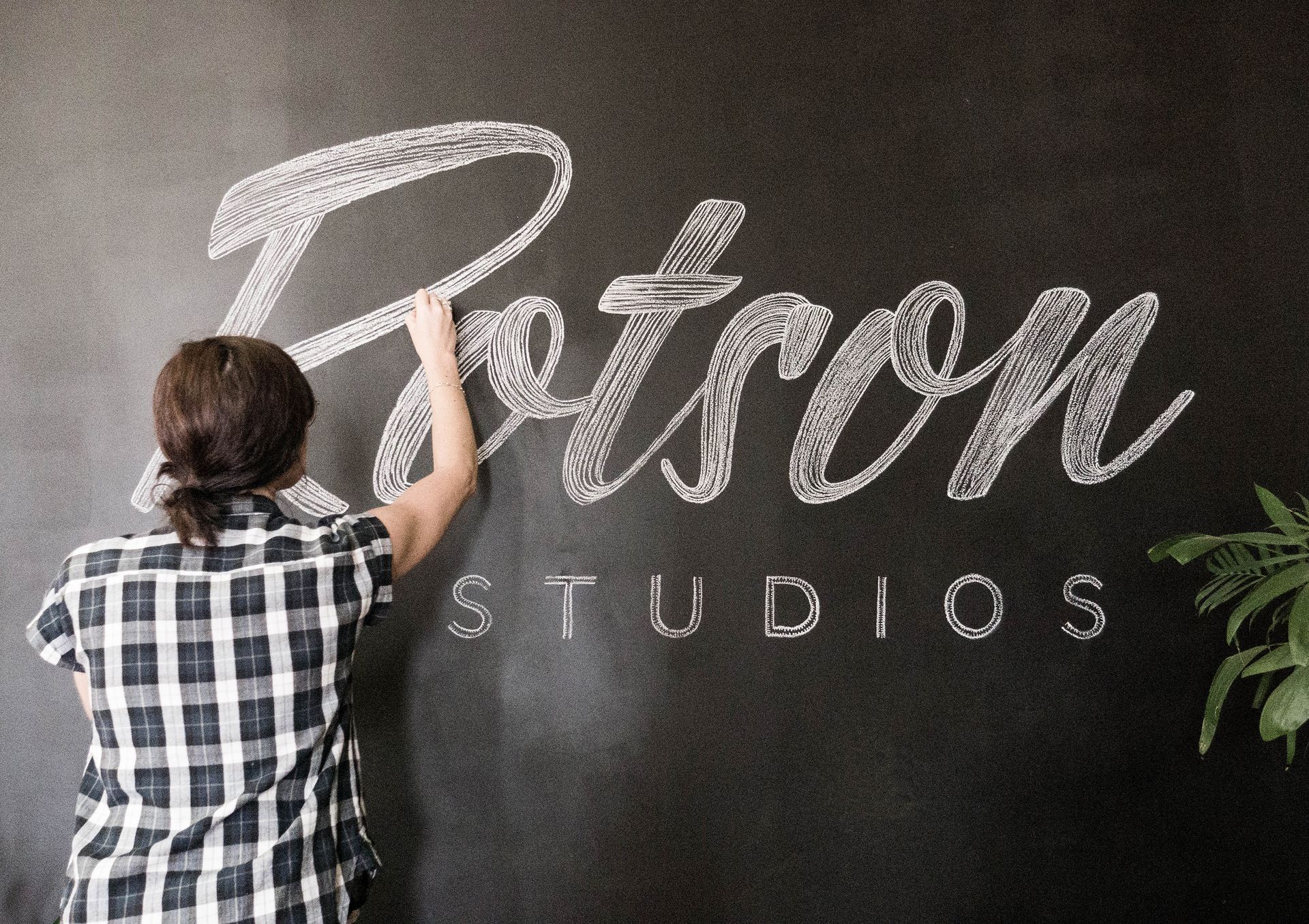 Maria Montes in plaid shirt lettering “Rotson Studios” on a black wall