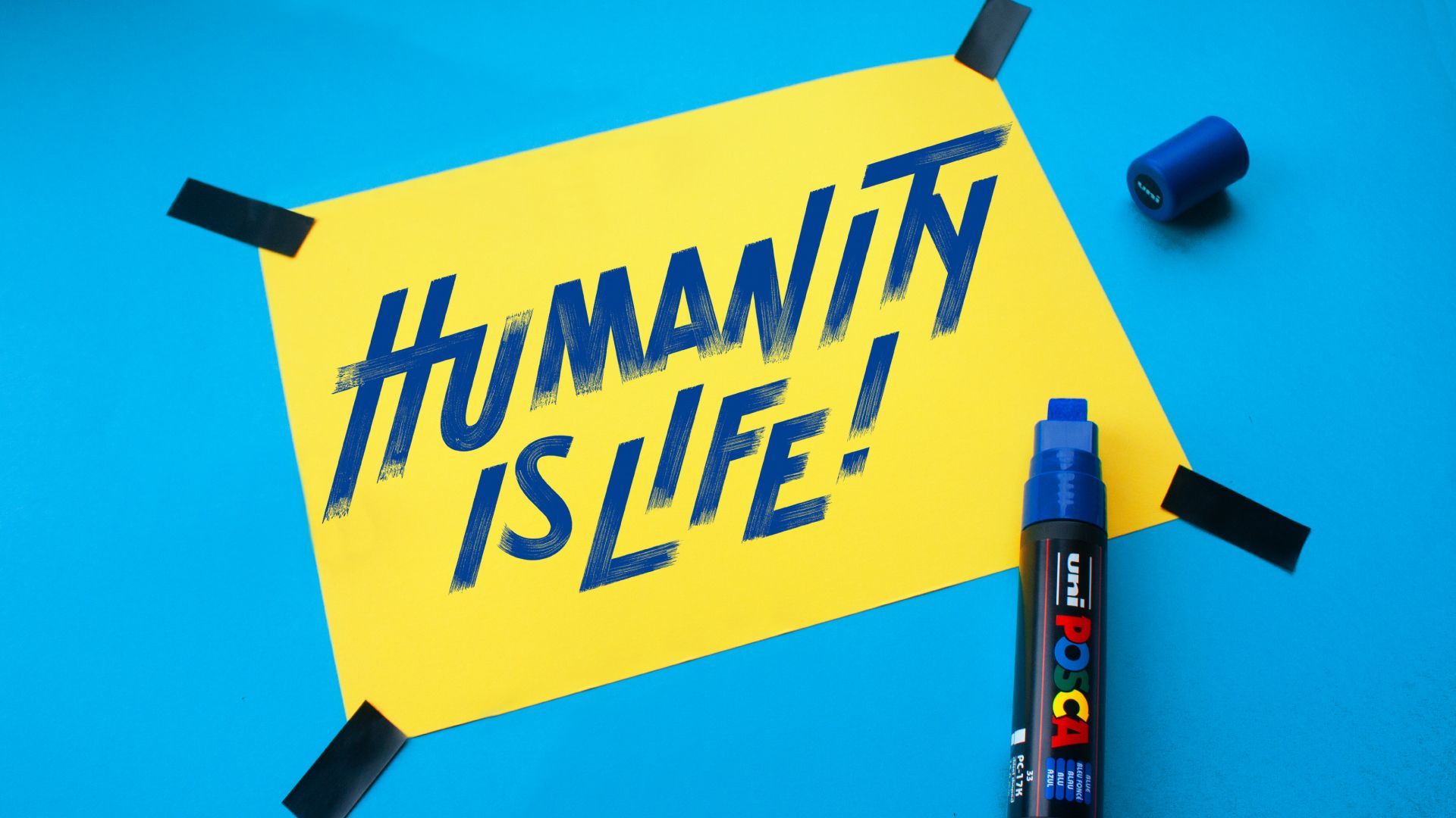 Yellow sticky note with “Humanity is life!” on blue background, taped beside a marker and cap