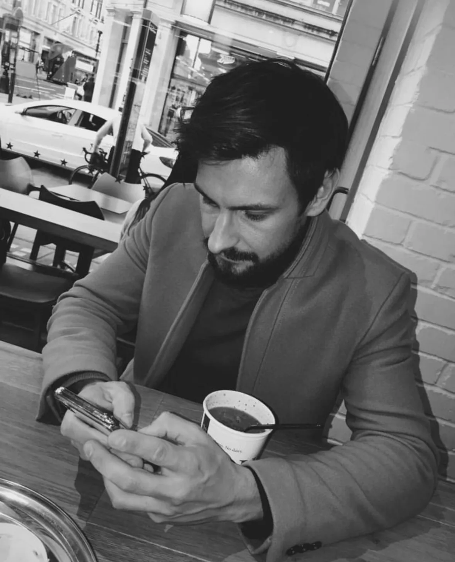 Man seated at an outdoor café, looking at his phone while holding a coffee cup.