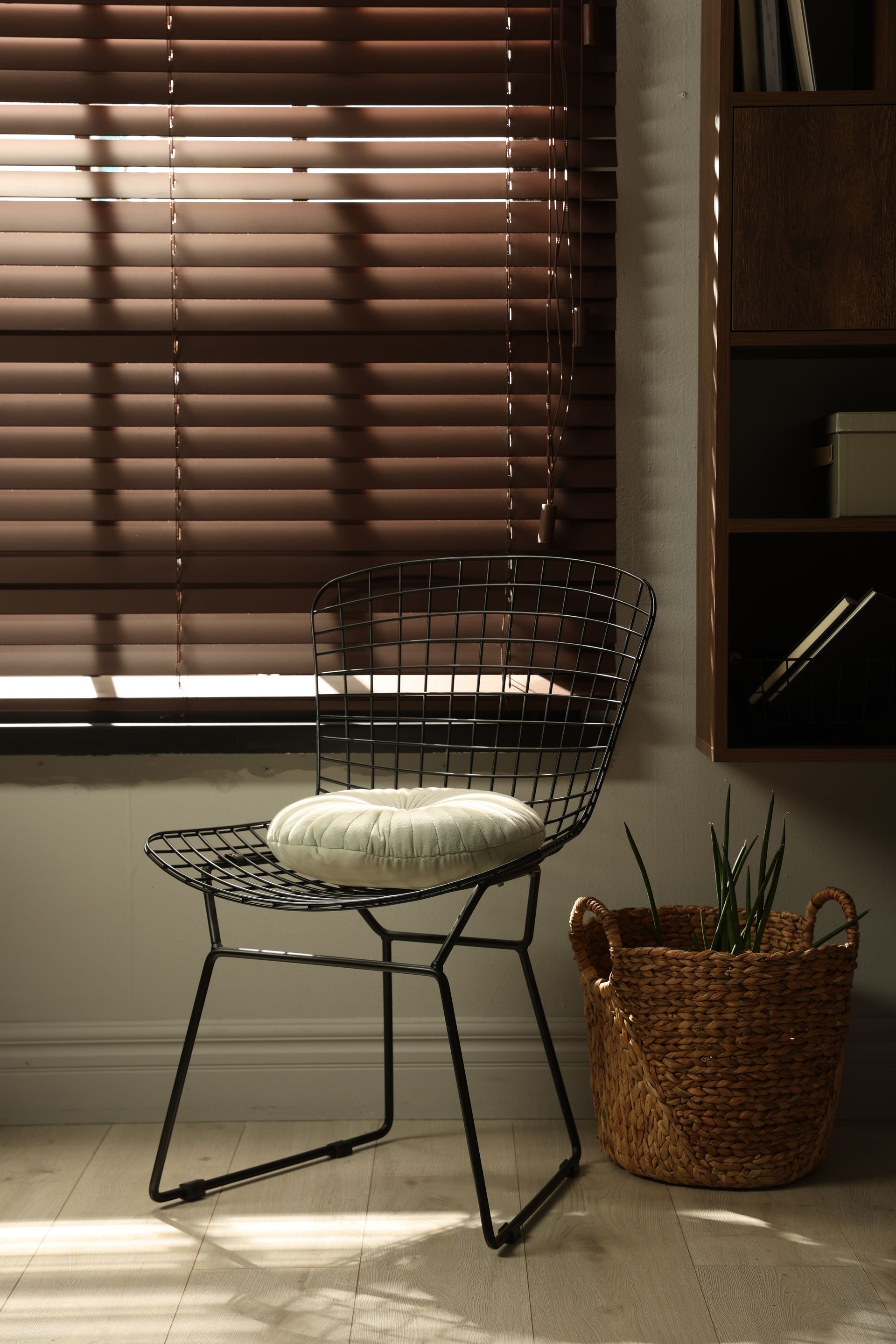 Wire chair with white cushion beside a wicker basket in a sunlit room with window blinds.