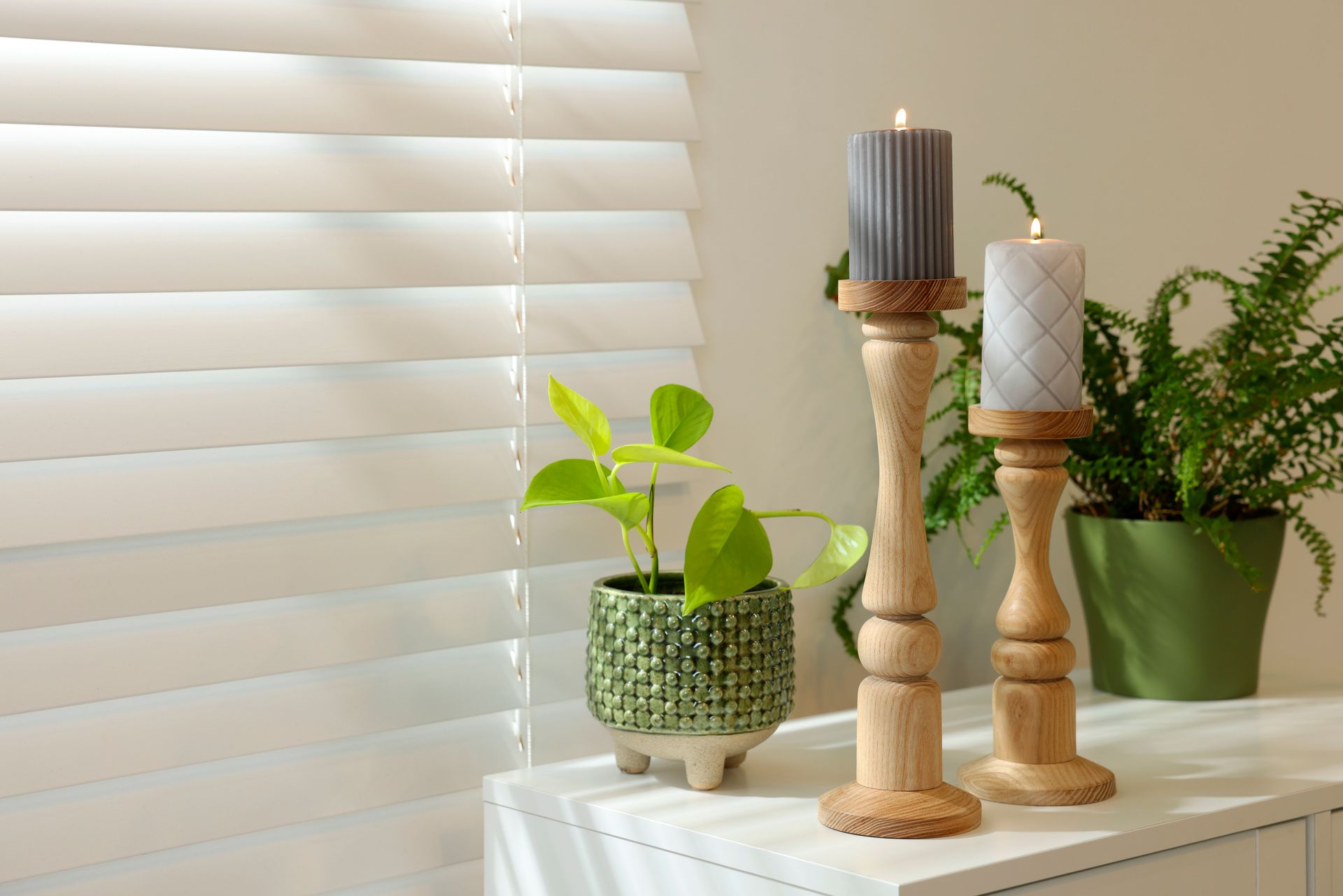 Candles and potted plants on a white table by a window with blinds