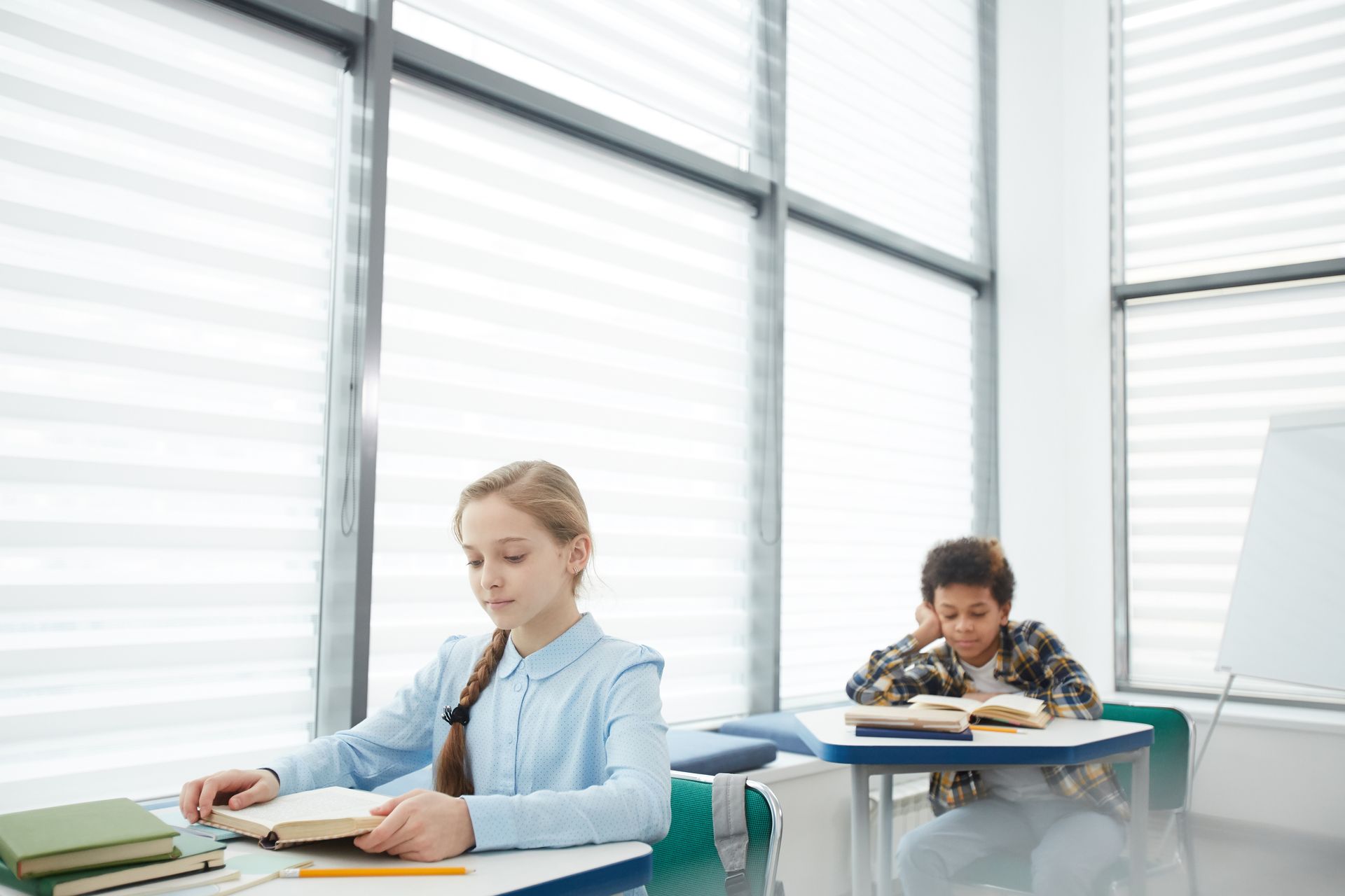 Two students working quietly at desks in a bright classroom by large windows