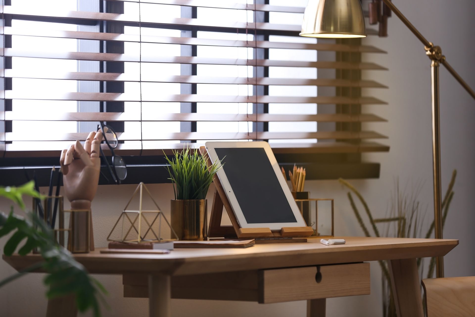 Desk by window with blinds, tablet on stand, lamp, plant, and pen holder in a cozy workspace.