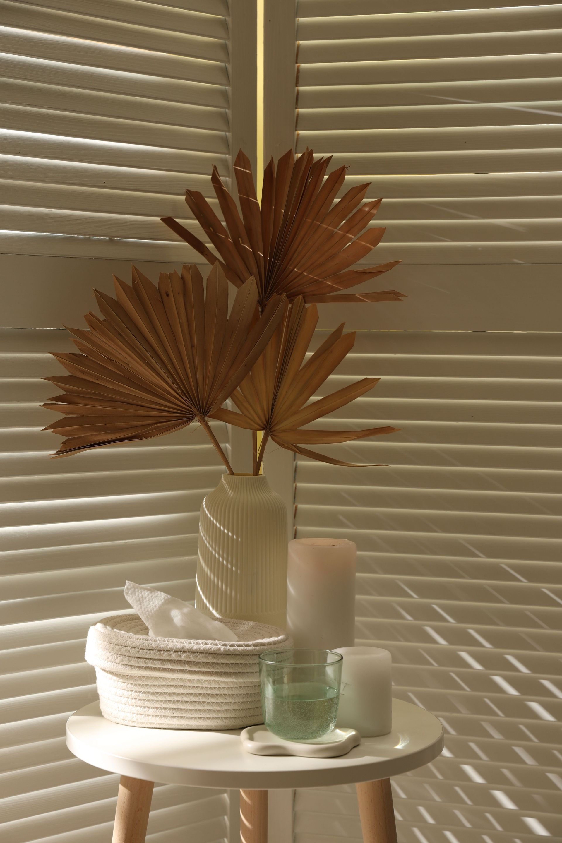 Decorative dried palm leaves in a vase on a white table beside candles and woven decor, against blinds.