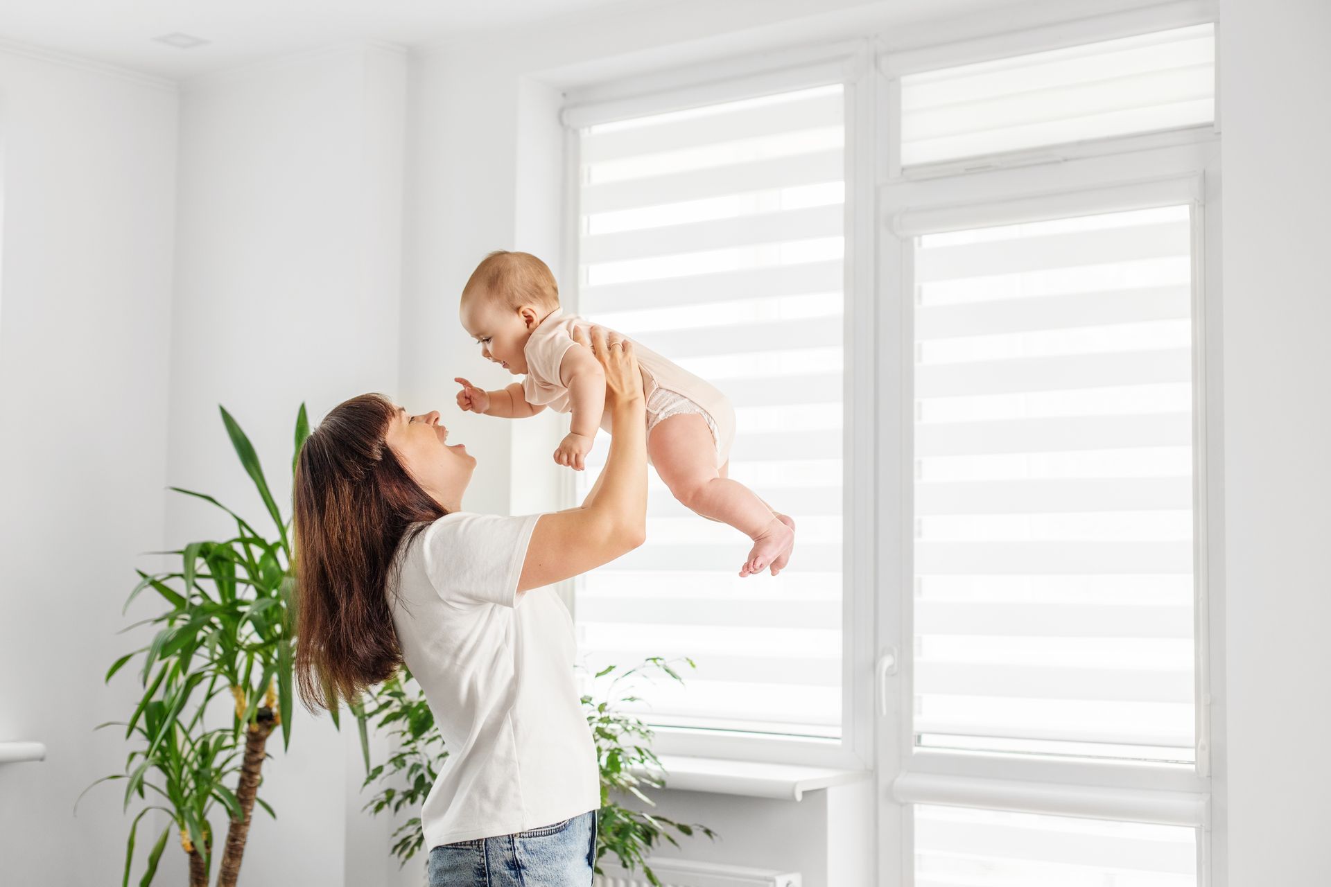 Woman lifts a baby near a bright window in a white room with a potted plant.