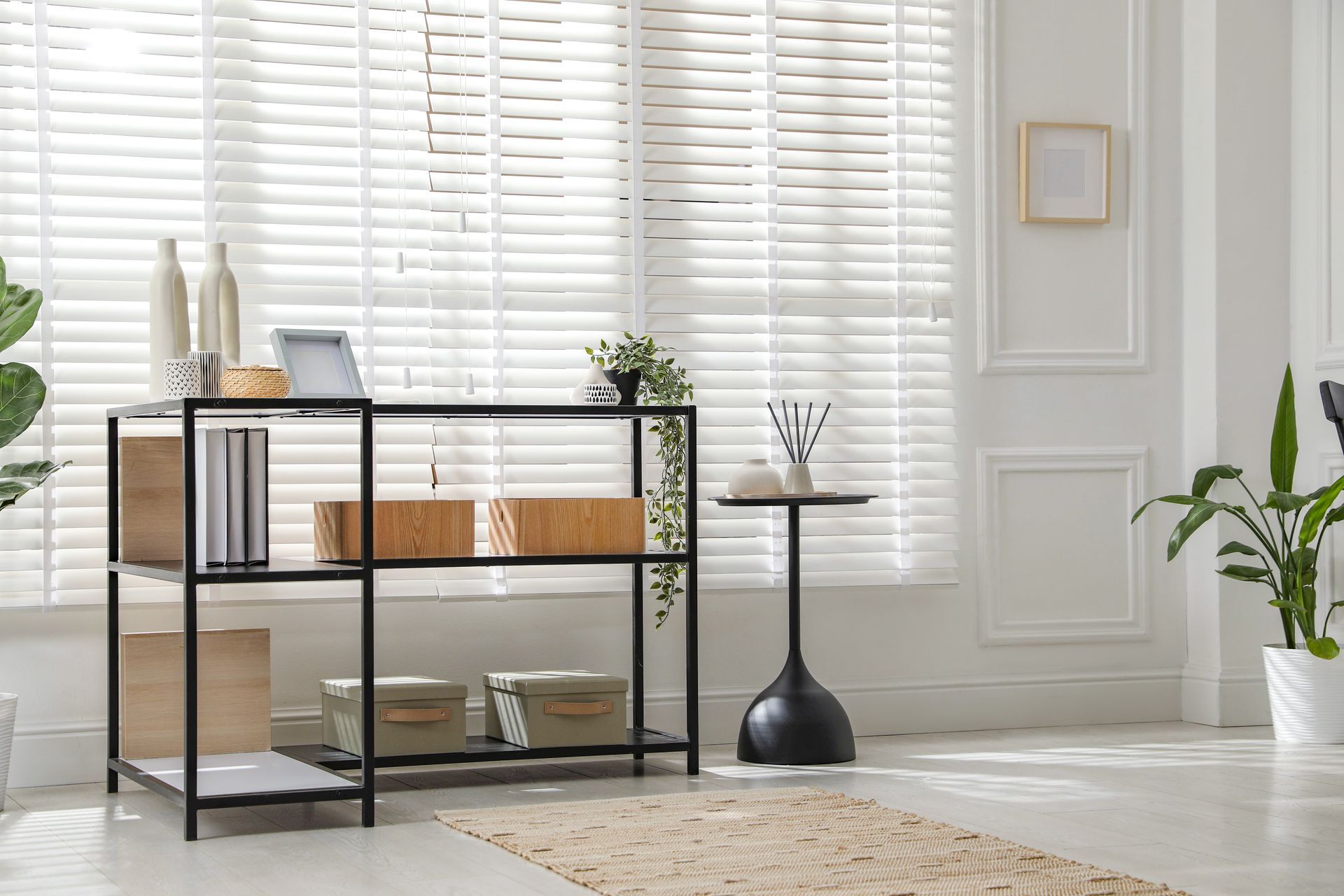 Minimalist living room with black shelving, white shutters, plants, and woven decor on a light rug.