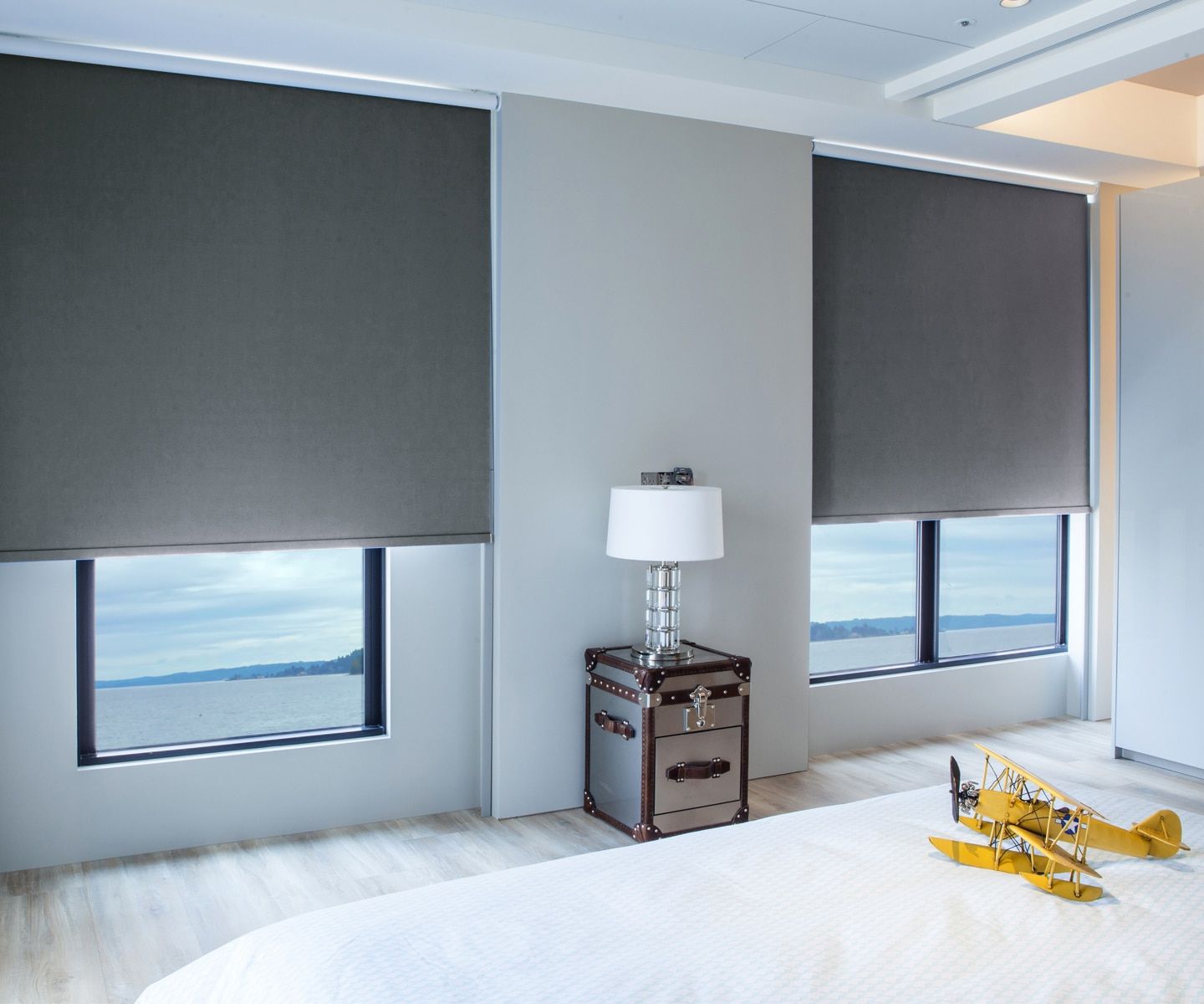 Modern bedroom with gray window shades, ocean view, white bed, and a small lamp on a bedside table