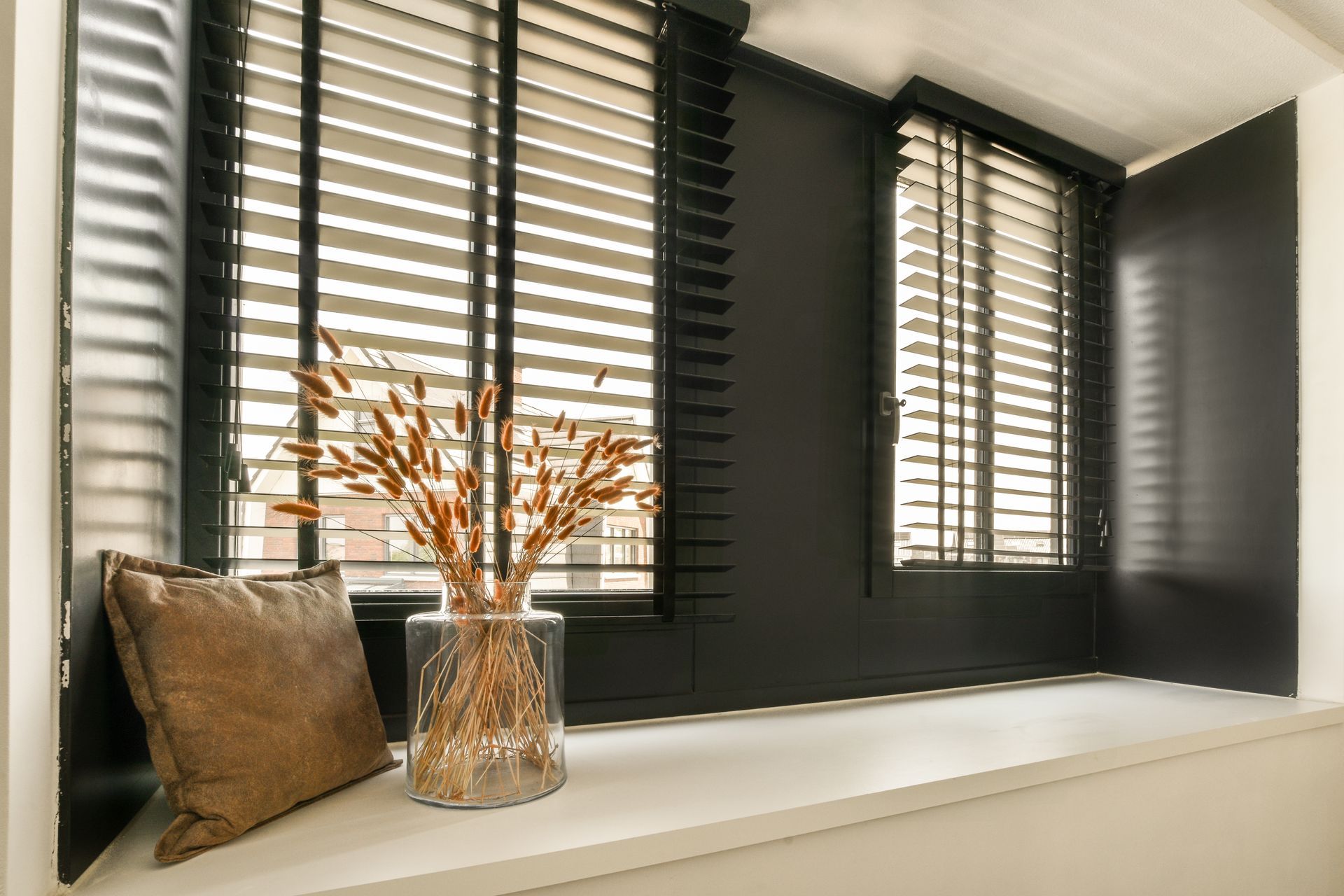 Black window seat with beige pillow, glass vase of dried stems, and white shutters by a window