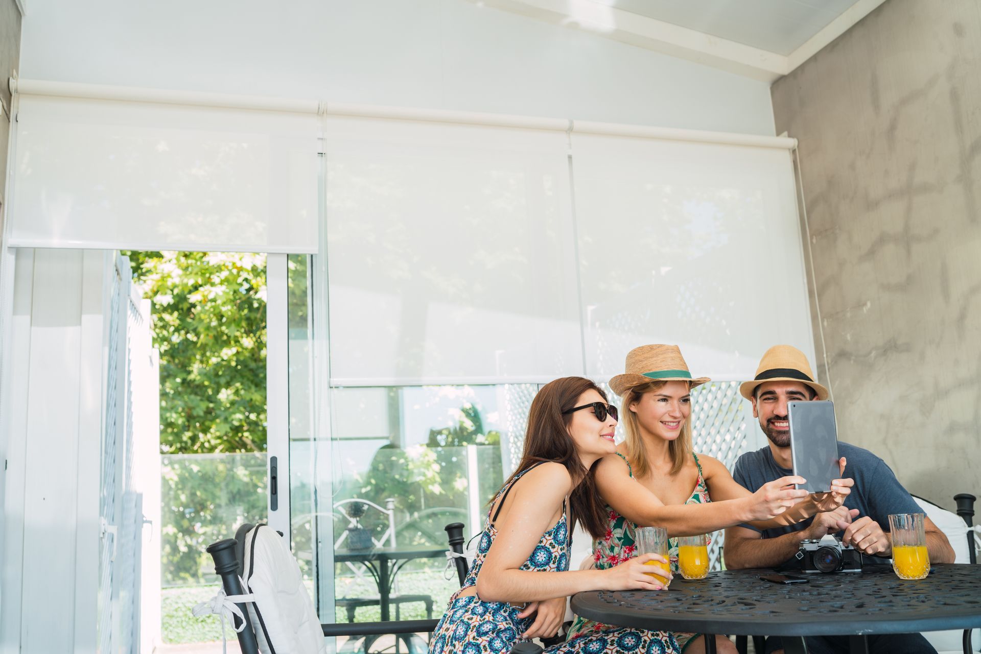 Three friends smiling at a table on a bright patio, sharing drinks and using a phone Three friends smiling at a table on a bright patio, sharing drinks and using a phone