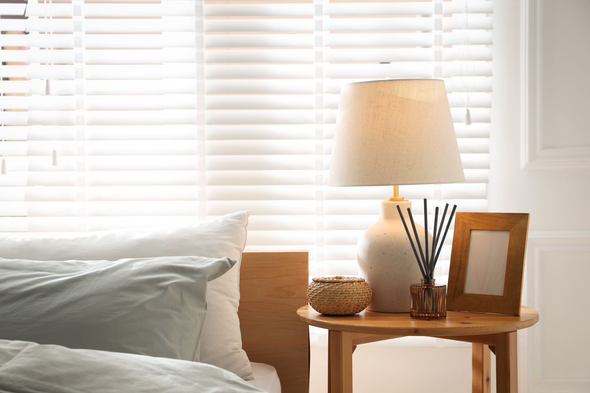 White bed by a sunlit window with blinds, next to a wooden side table, lamp, and framed photo