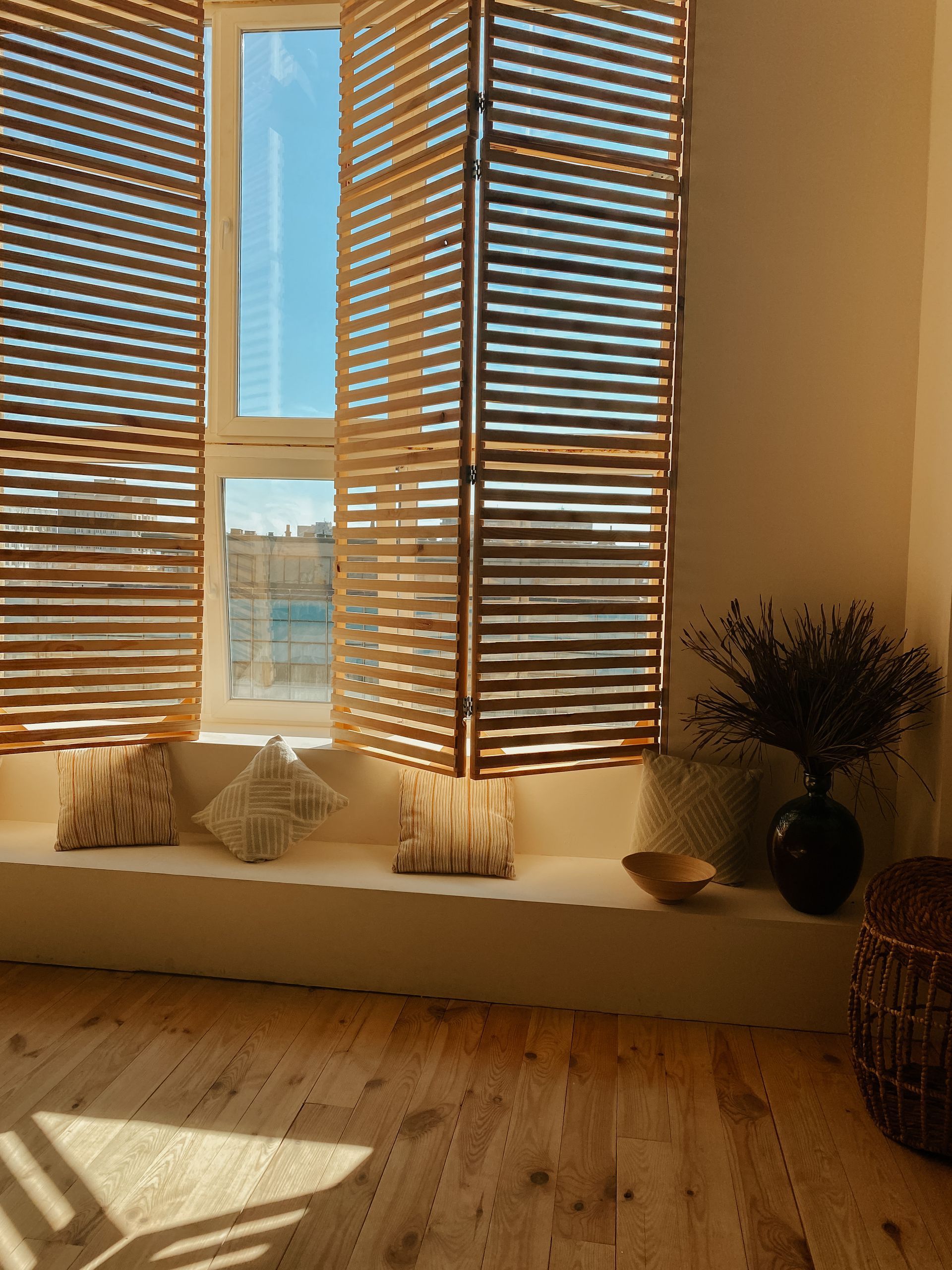 Sunlit room with wooden blinds, window seat cushions, and a vase of dried flowers on the right.