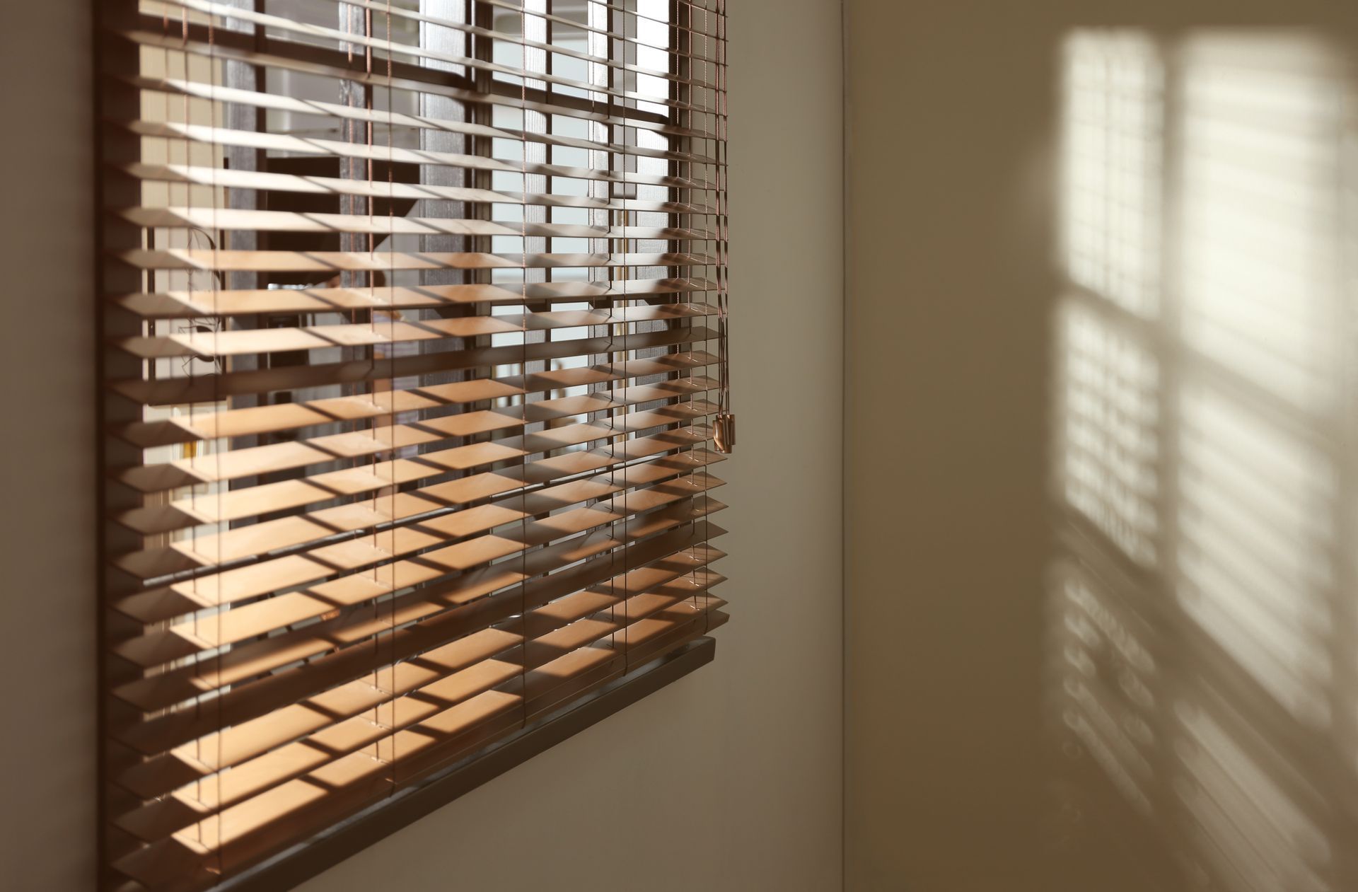 Sunlight through wooden blinds casts striped shadows on a beige wall. Sunlight through wooden blinds casts striped shadows on a beige wall.