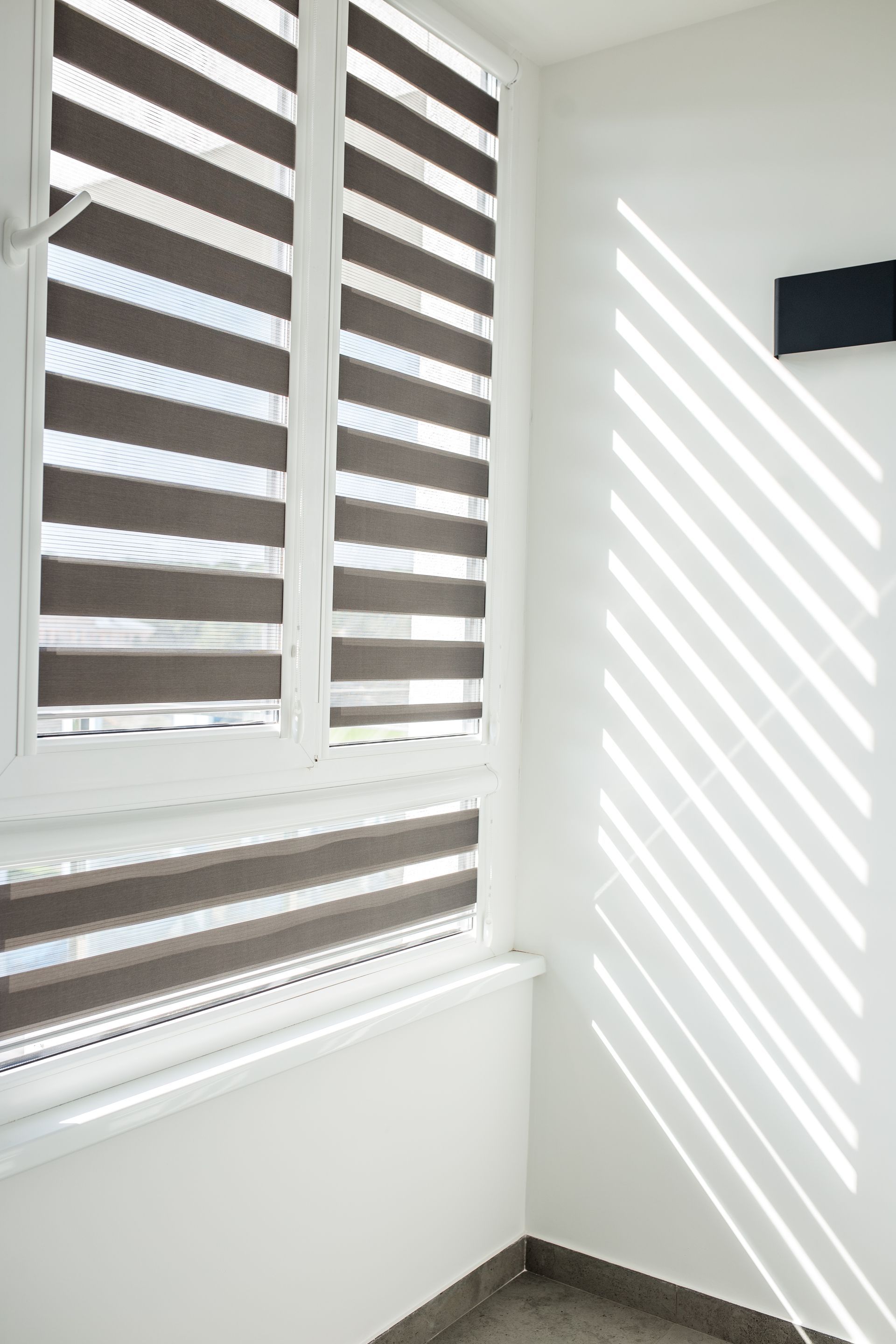 White blinds with sunlight casting striped shadows on a white wall and window ledge.