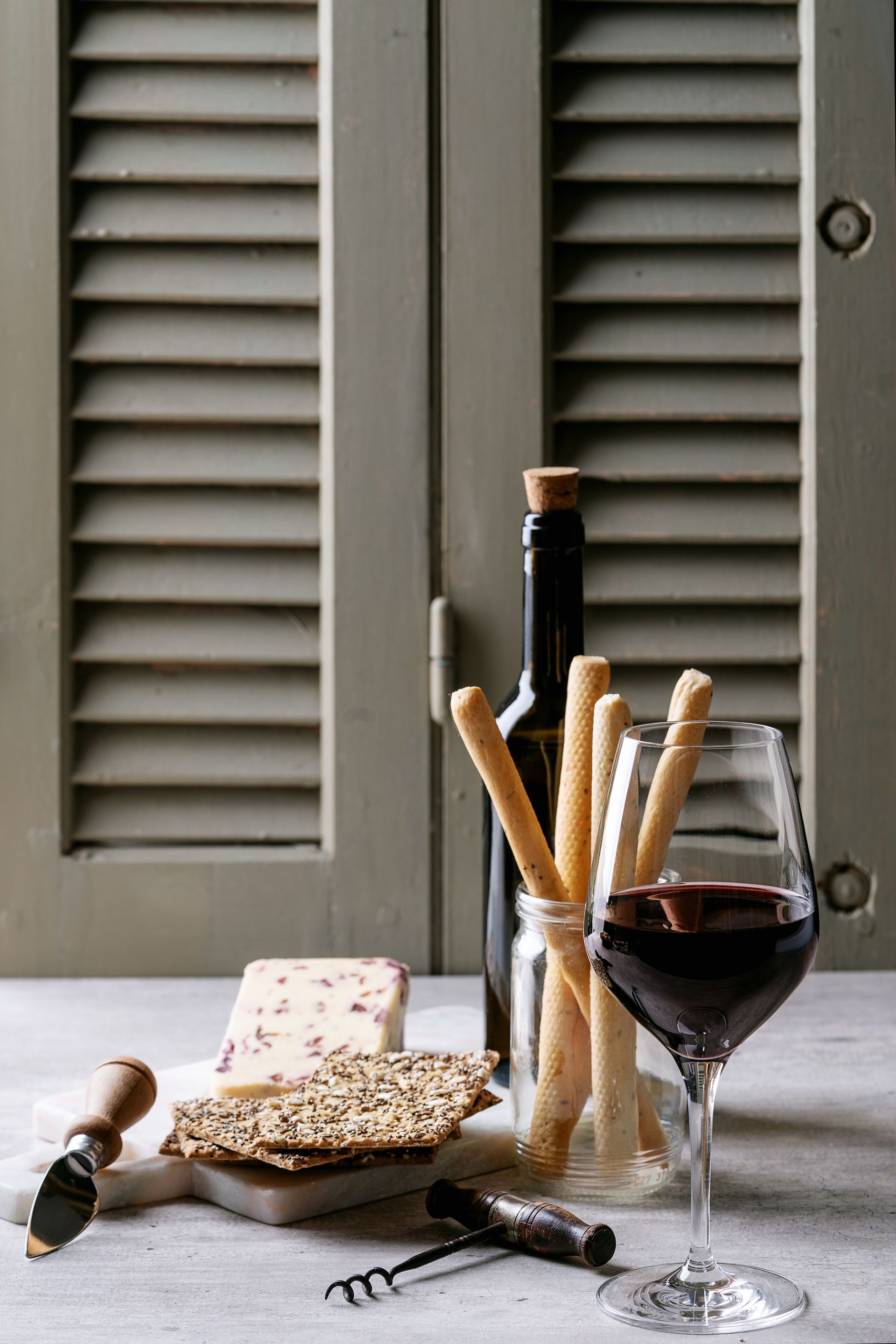 Wine glass, breadsticks, cheese, and a bottle on a rustic table by gray shutters.
