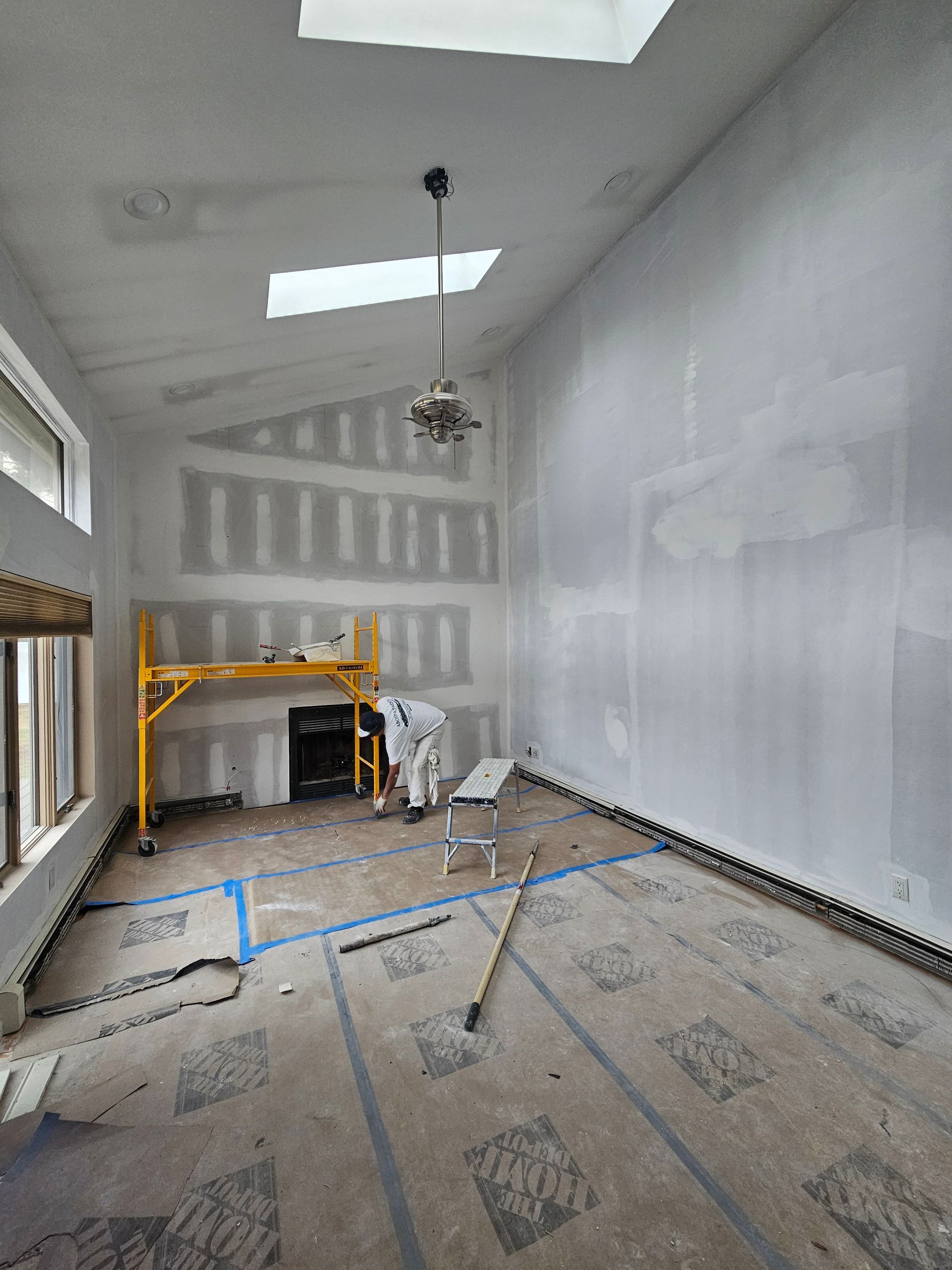 Man in gloves using a drill to install drywall on a ceiling with metal framing.
