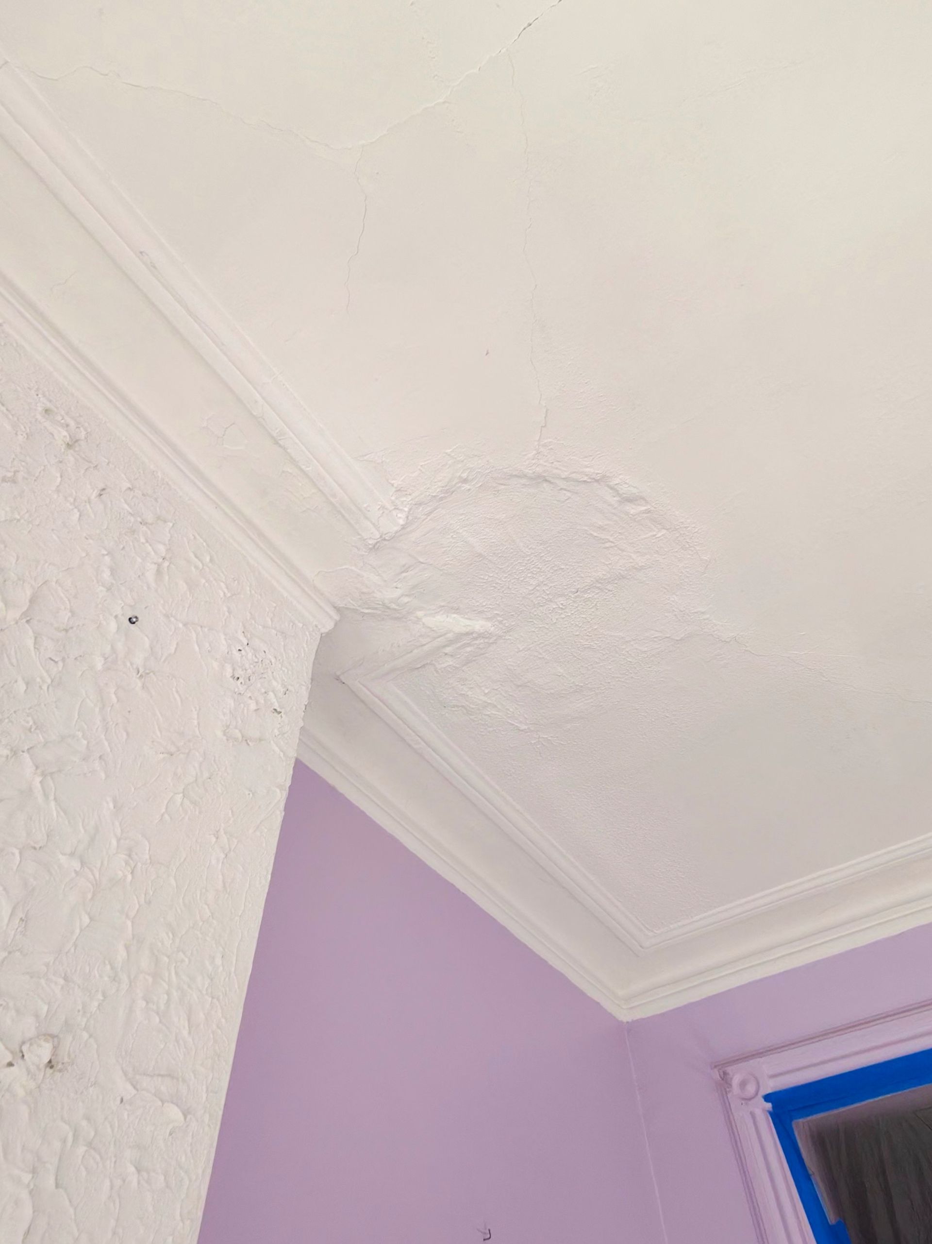 Construction worker painting a wall with a roller; wearing an orange hard hat, overalls, and a plaid shirt.