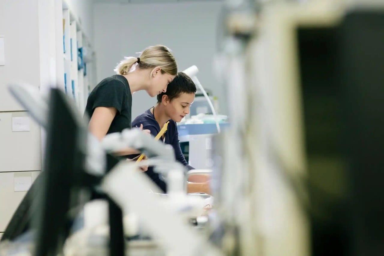 Una donna si china per aiutare una persona che lavora con le attrezzature in un laboratorio.