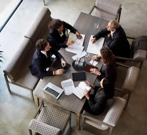Un uomo in giacca e cravatta gesticola durante una riunione a un tavolo con altre persone.