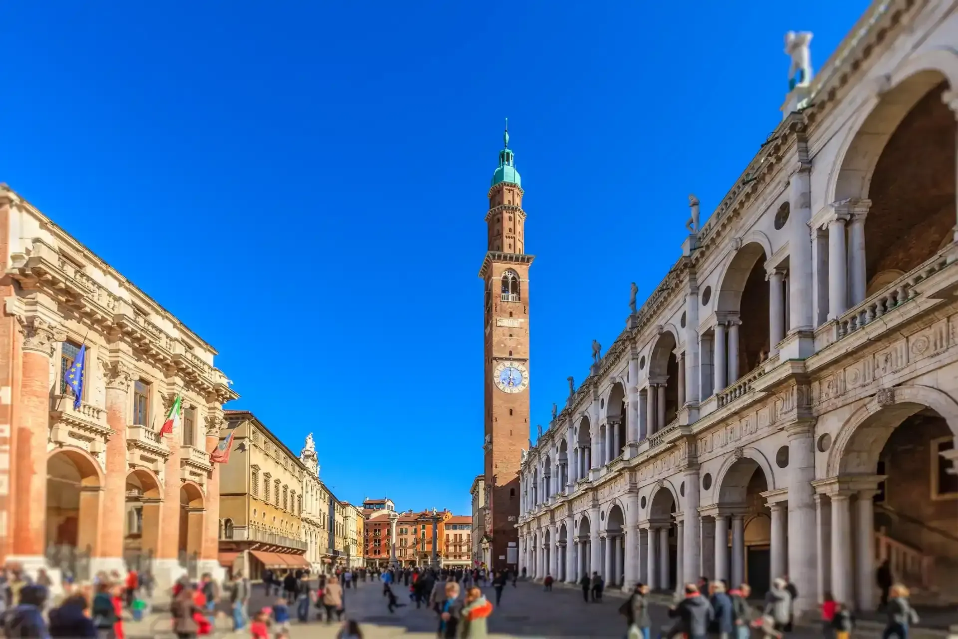 Piazza dei Signori a Vicenza, in Italia, con la torre dell'orologio e i portici, brulicante di gente sotto un cielo azzurro brillante.