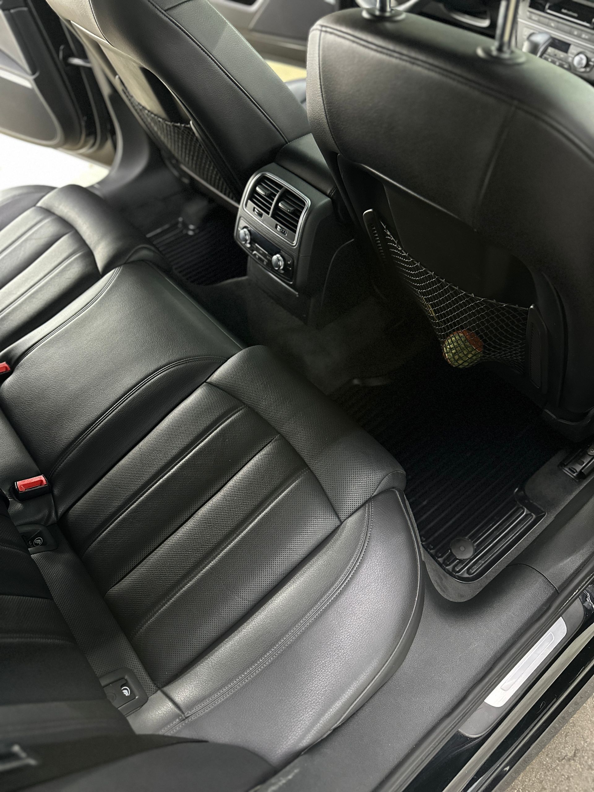 The interior backseat of a vehicle featuring black perforated leather seats, a center console air vent, and floor mats.