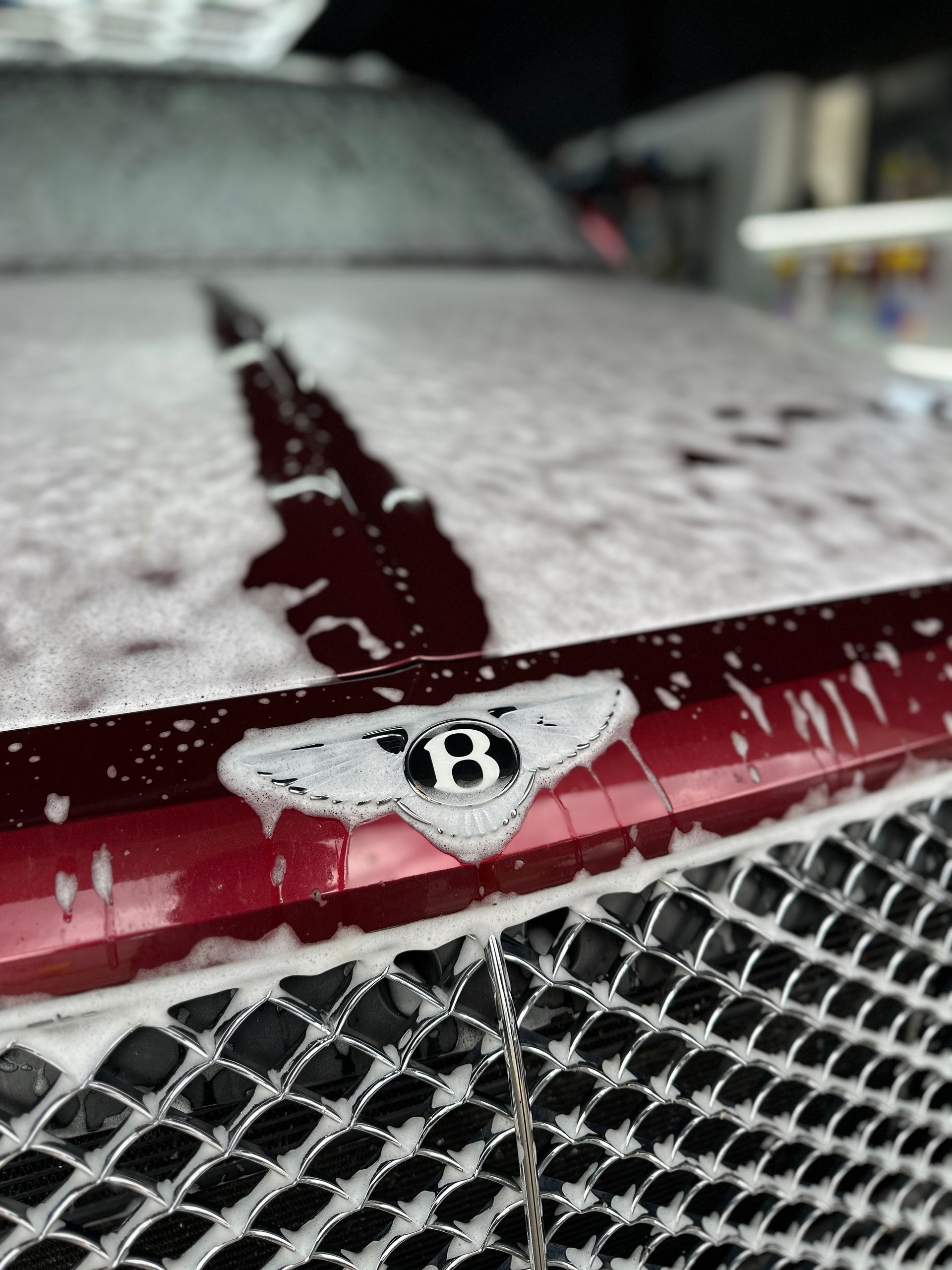 A close-up of a maroon Bentley car hood and chrome grille covered in thick white soap suds during a car wash.
