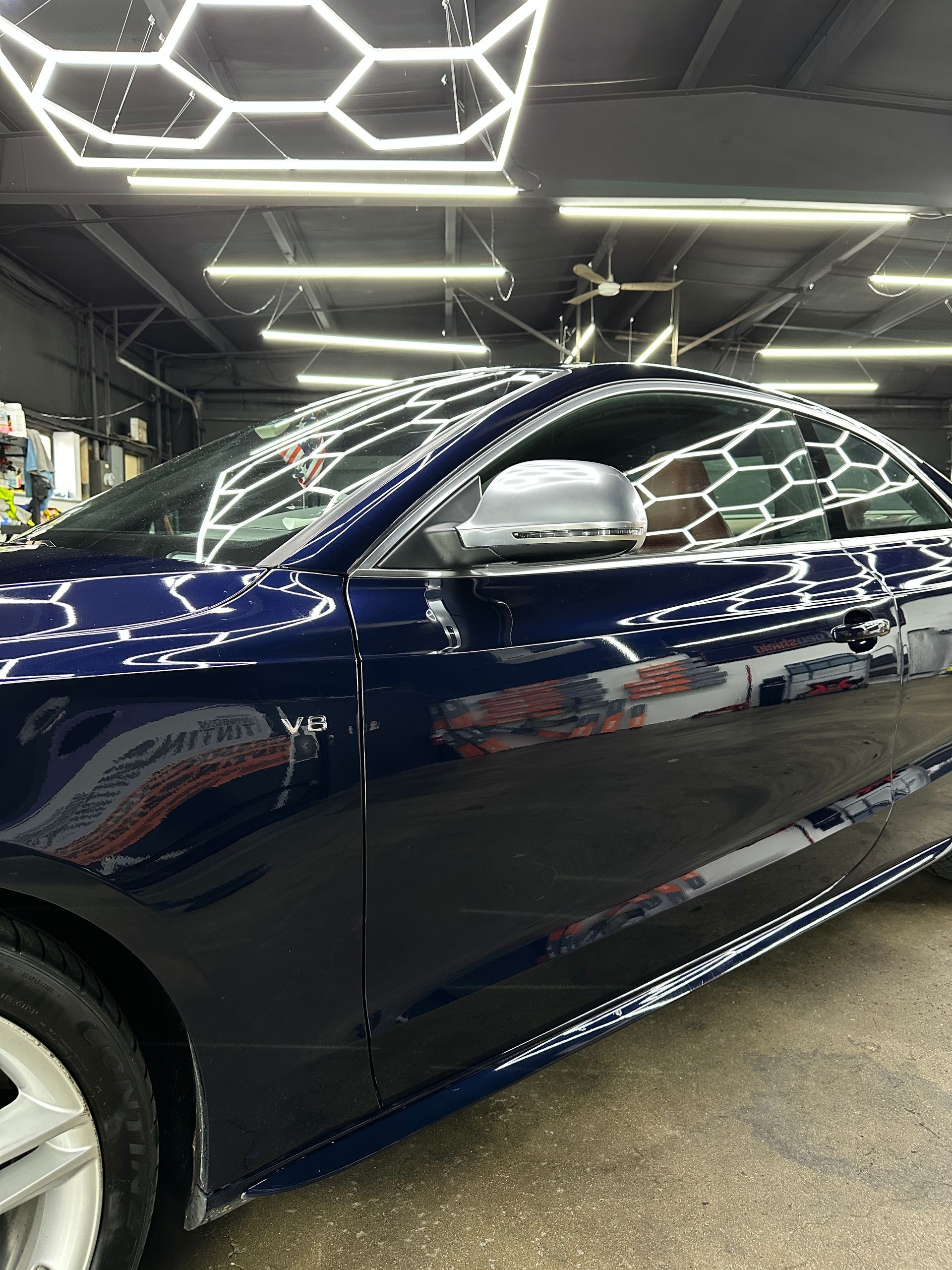 A side profile of a dark blue luxury car parked in a brightly lit workshop with hexagonal ceiling lights.