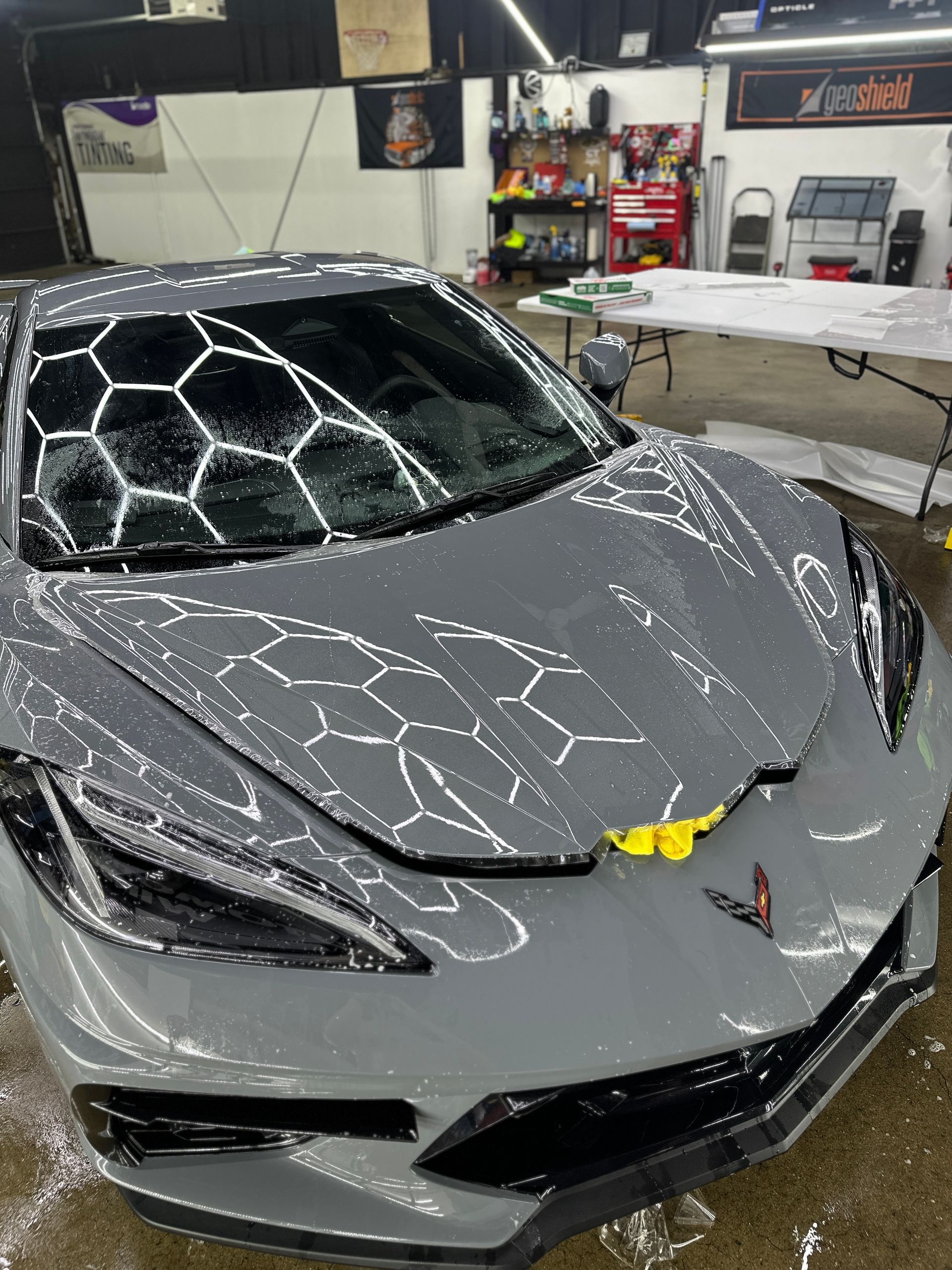 A gray Corvette in a garage covered in soap suds forming a honeycomb pattern on its windshield and hood.