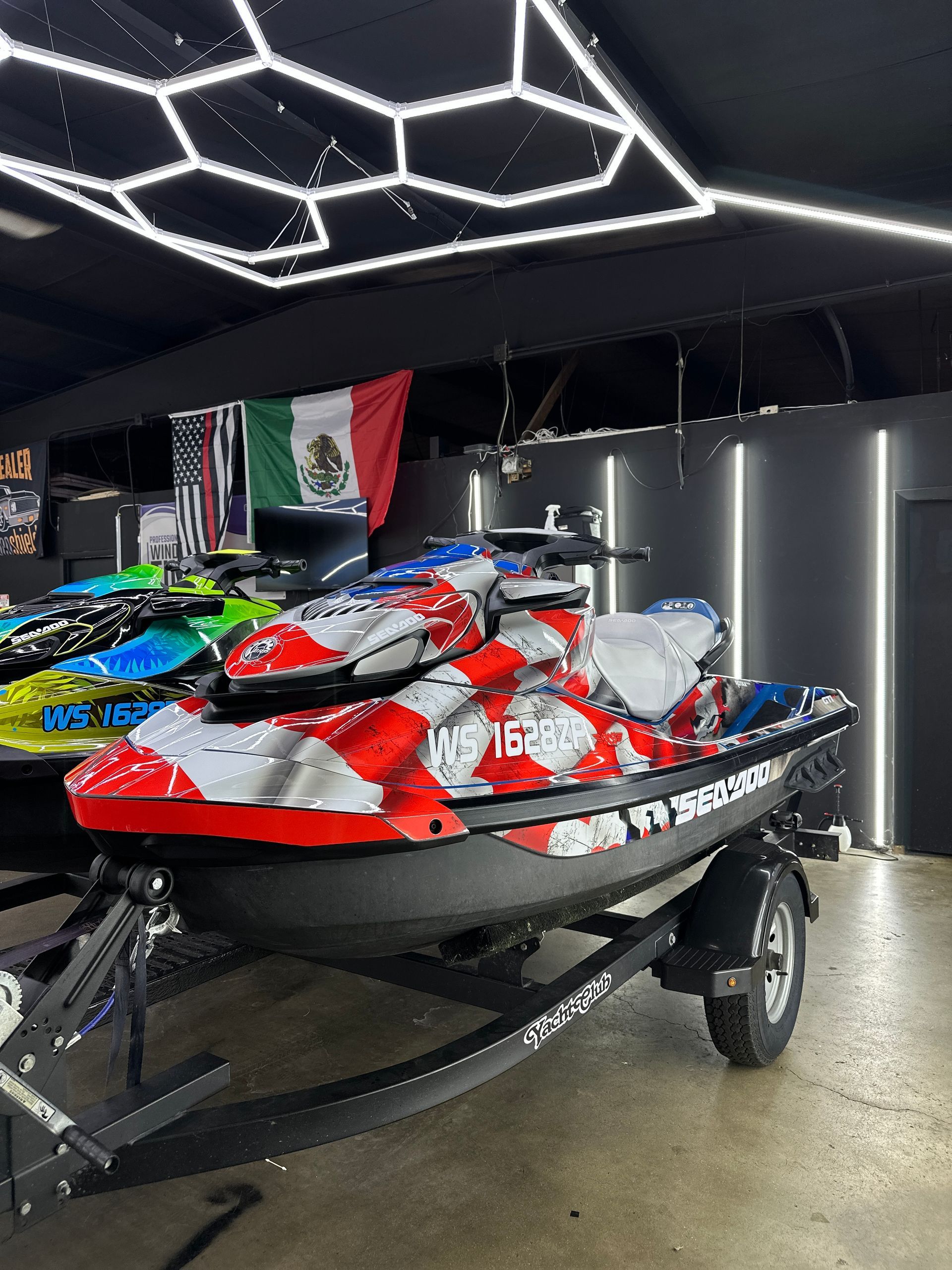 A red, white, and blue jet ski on a trailer inside a garage with bright geometric ceiling lights and a Mexican flag.