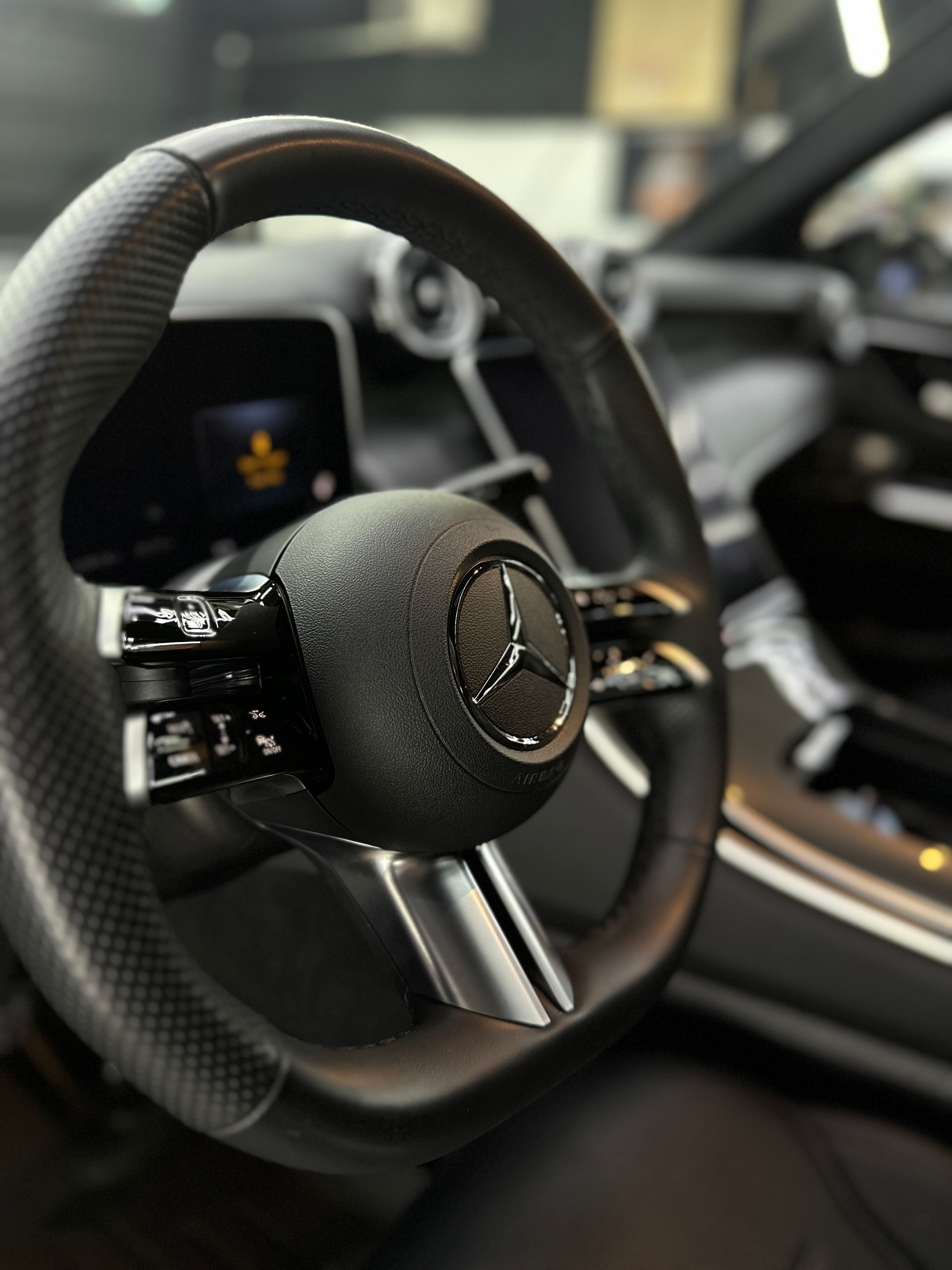 Close-up interior view of a black Mercedes-Benz steering wheel featuring the brand logo and modern dashboard displays.