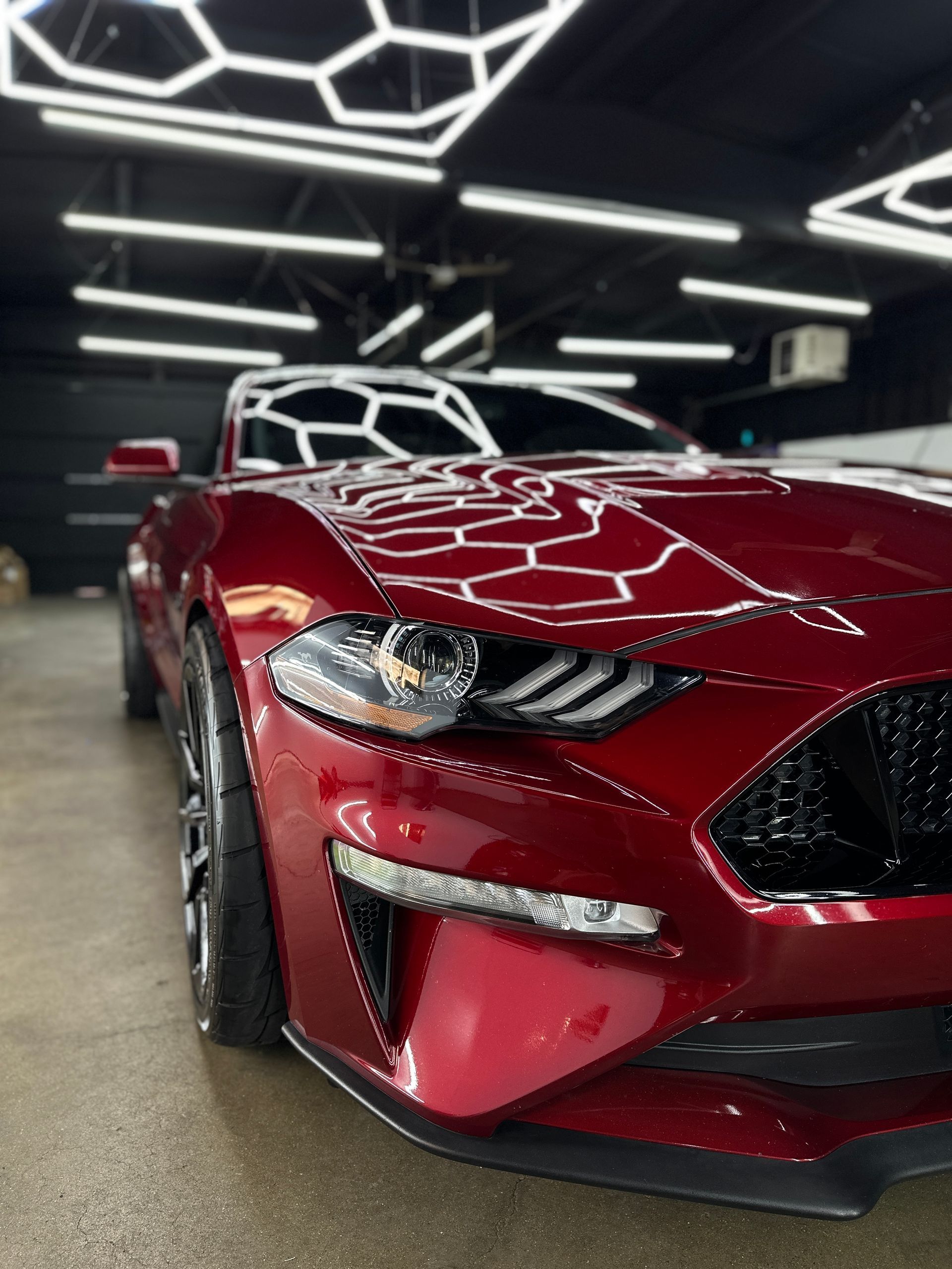 A glossy, dark red Ford Mustang parked in a well-lit garage with geometric ceiling lights reflecting on its hood.