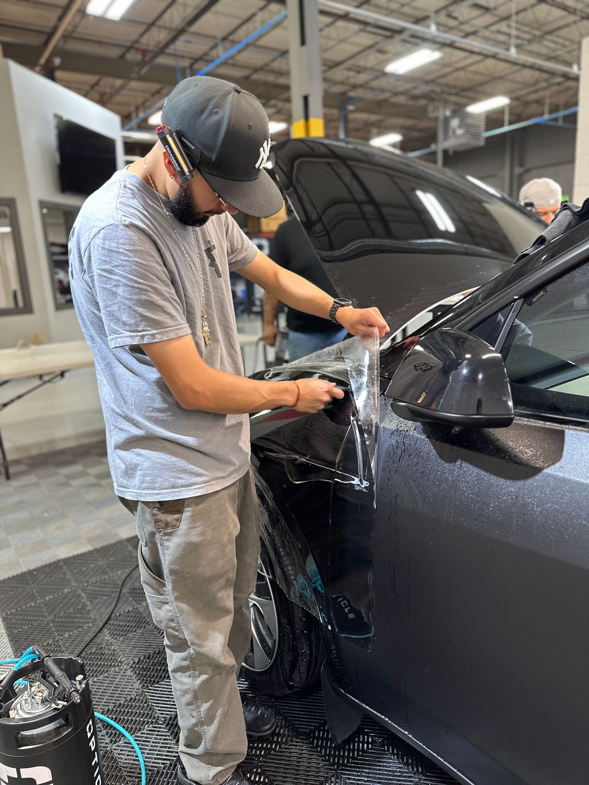A person wearing a hat applies a clear protective film to the side panel of a dark vehicle inside a workshop.