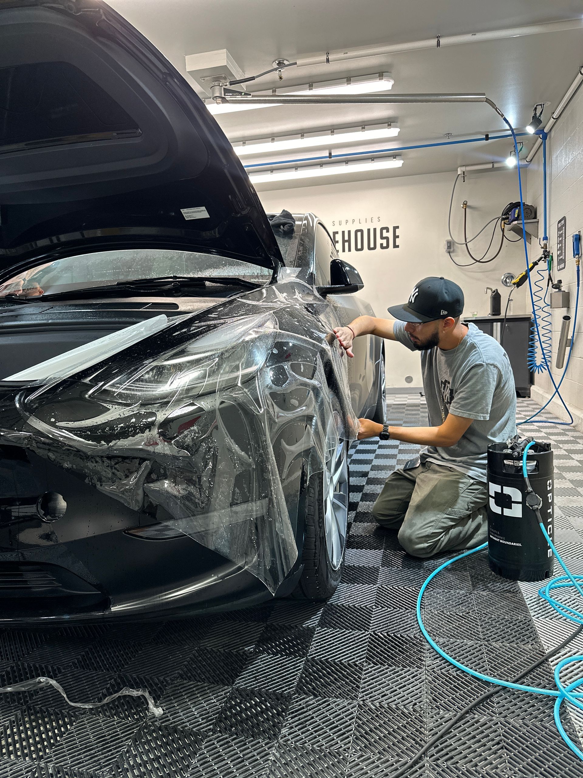 A technician in a garage applies a clear protective film to the front fender of a dark car.