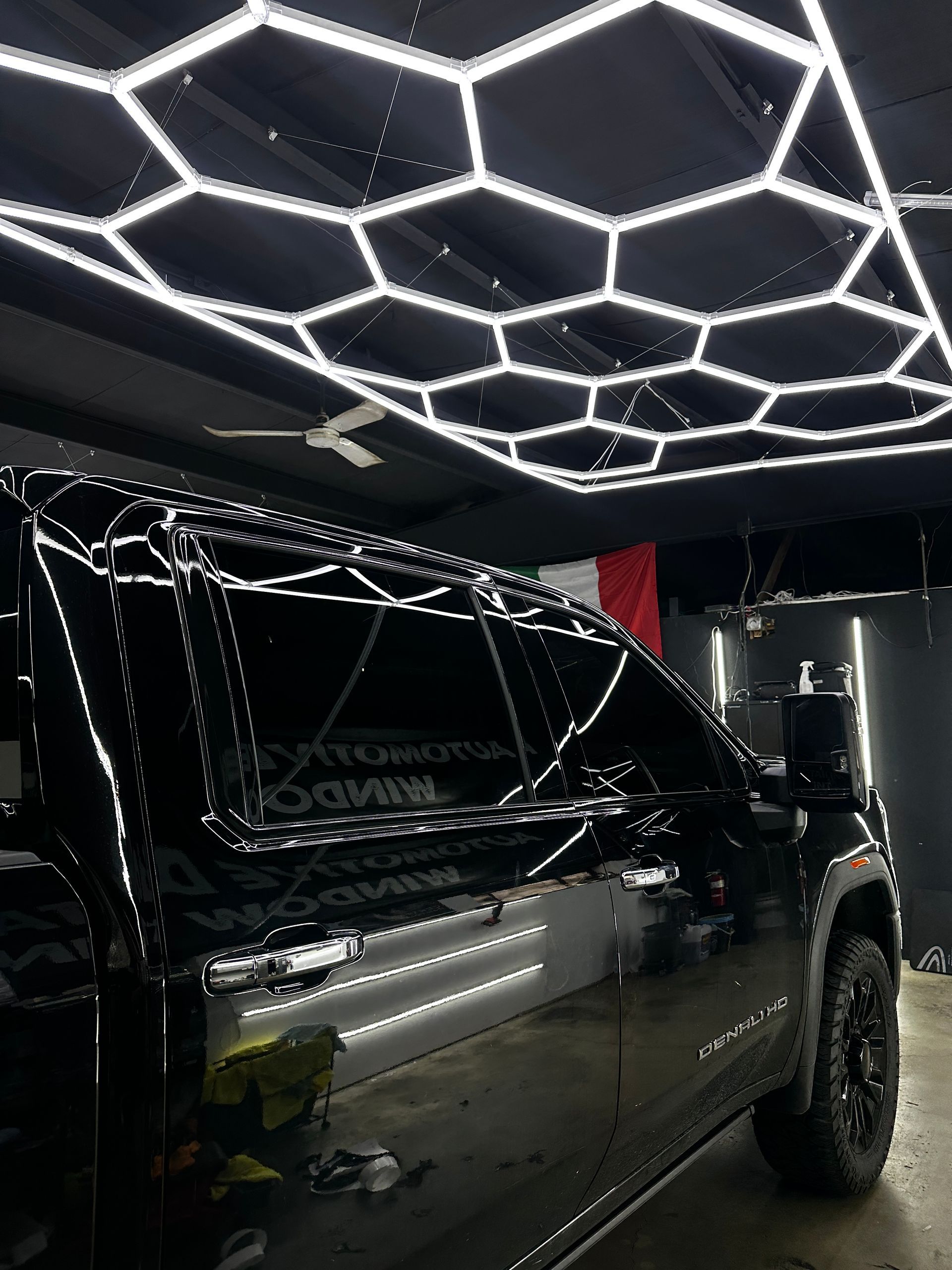 A side view of a glossy black pickup truck parked in a shop with a hexagonal LED ceiling light grid reflecting on its body.