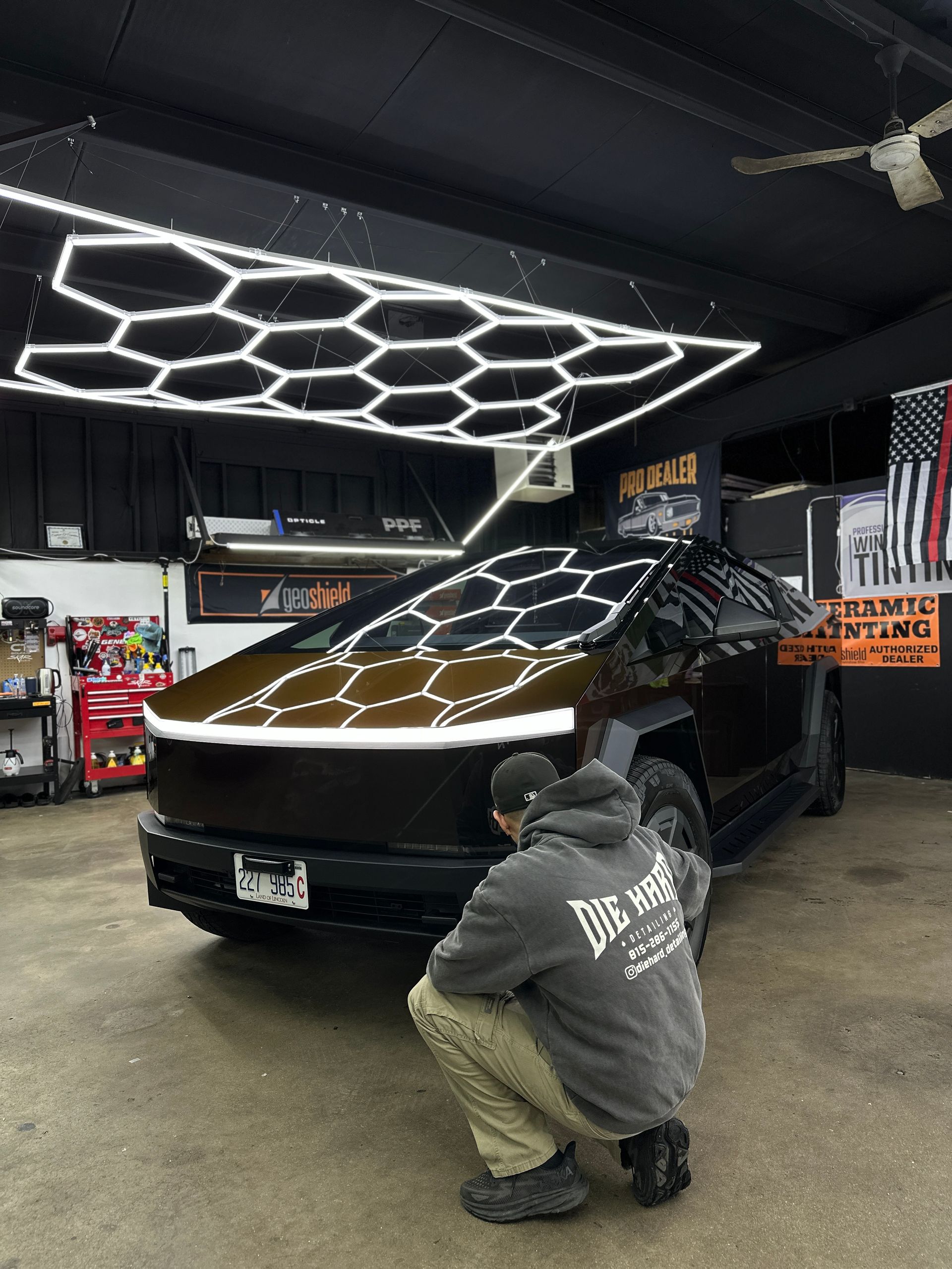 A person in a gray hoodie kneels by a metallic bronze Tesla Cybertruck in a garage with hexagonal ceiling lighting.