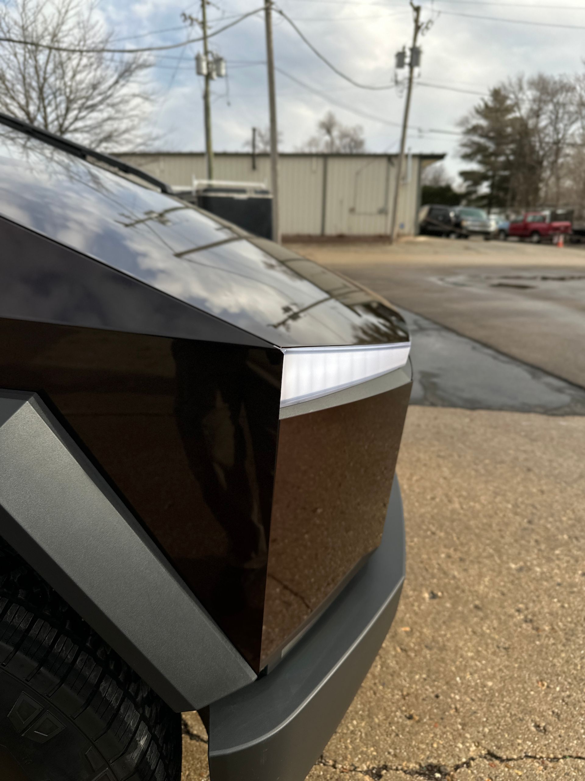 Close-up of a Tesla Cybertruck's angular stainless steel hood, headlight bar, and dark grey bumper parked outdoors.