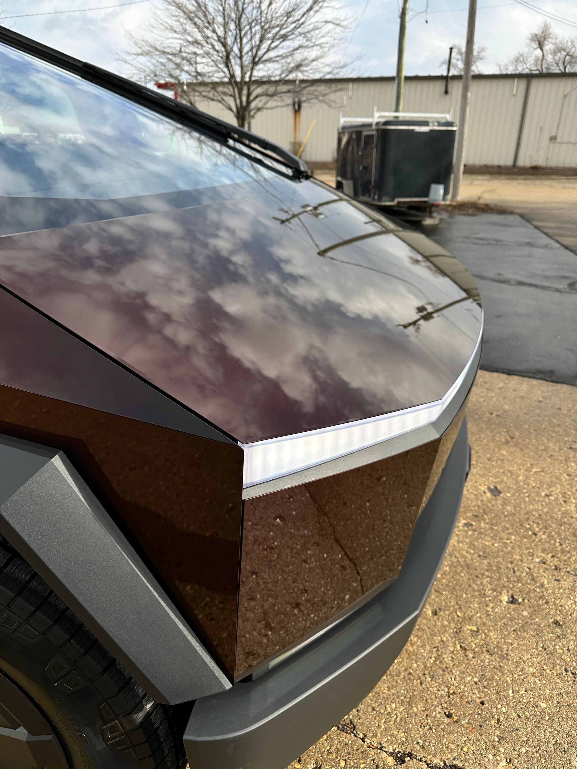 Close-up of a maroon-wrapped Tesla Cybertruck’s angular front hood, windshield, and LED headlight bar outdoors.