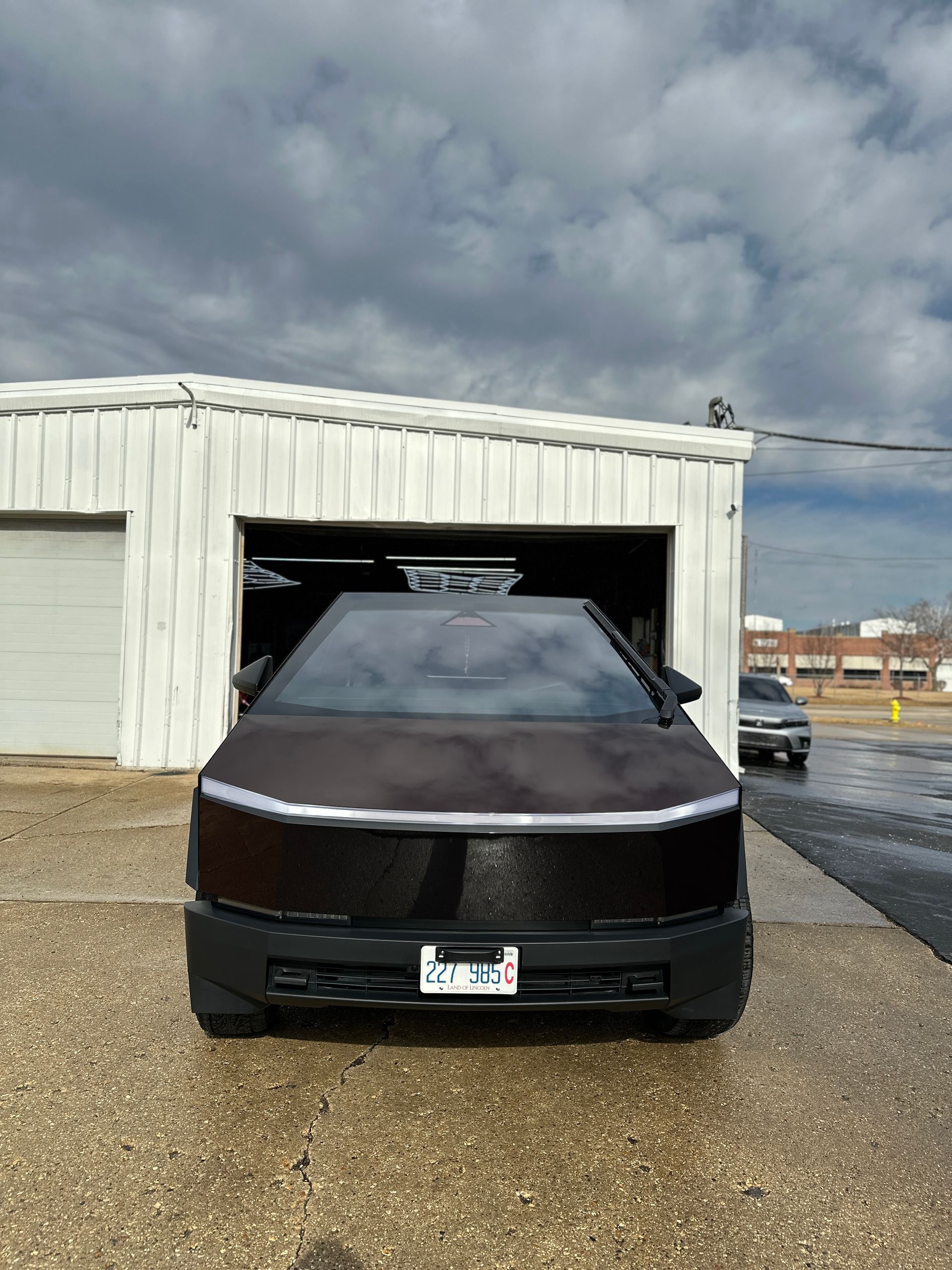 A dark Tesla Cybertruck with a front light bar parked in front of a white garage door under a cloudy sky.