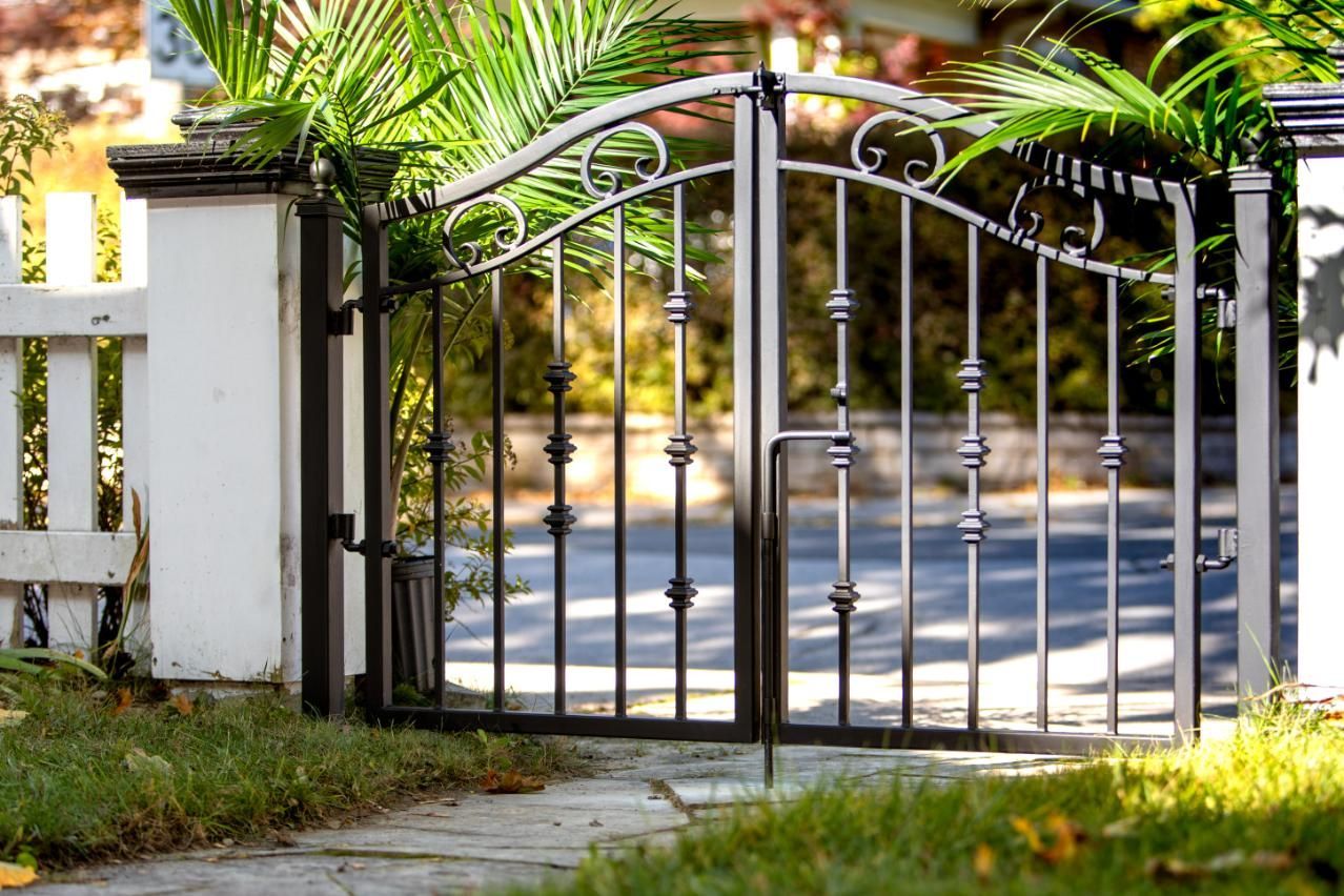 A wrought iron gate is open in front of a white fence.