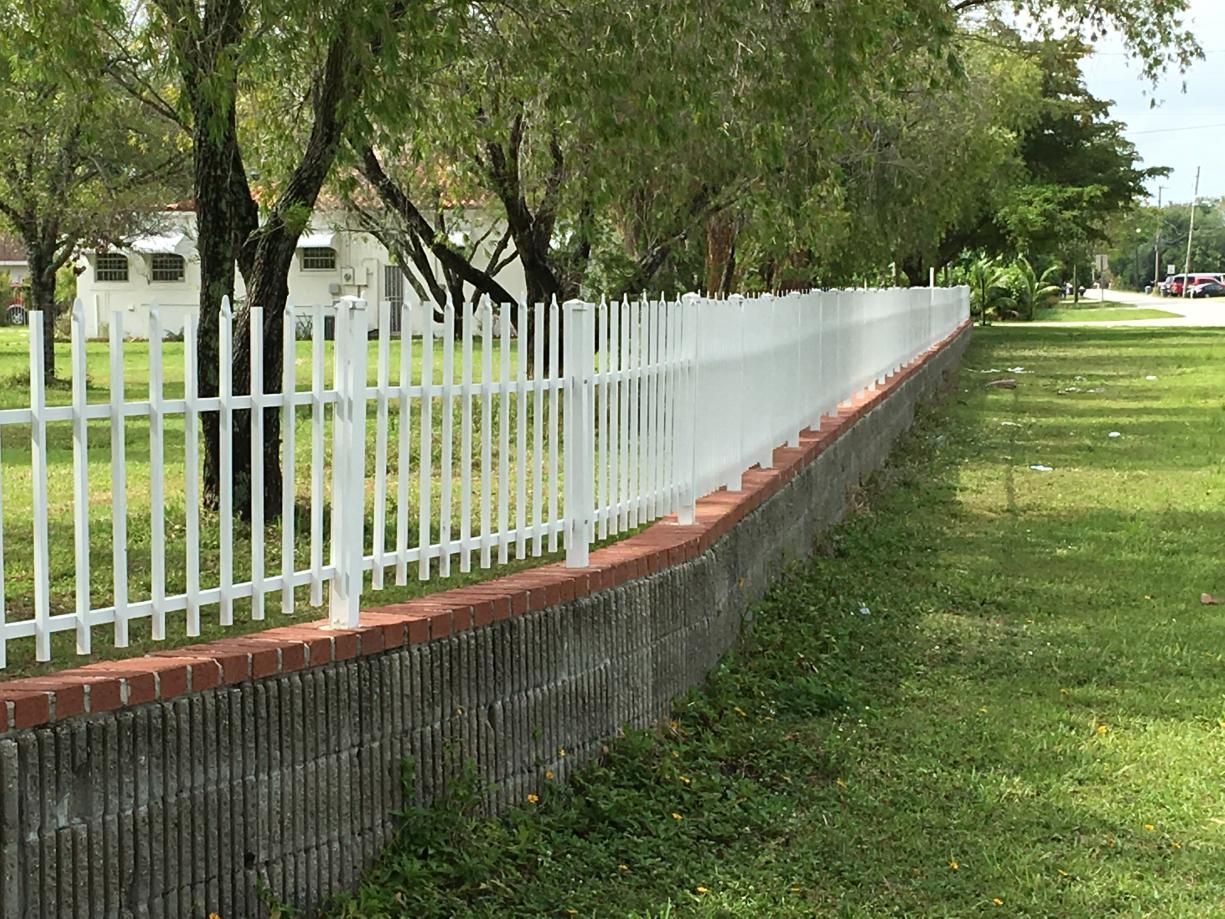A white picket fence surrounds a brick wall in a yard.